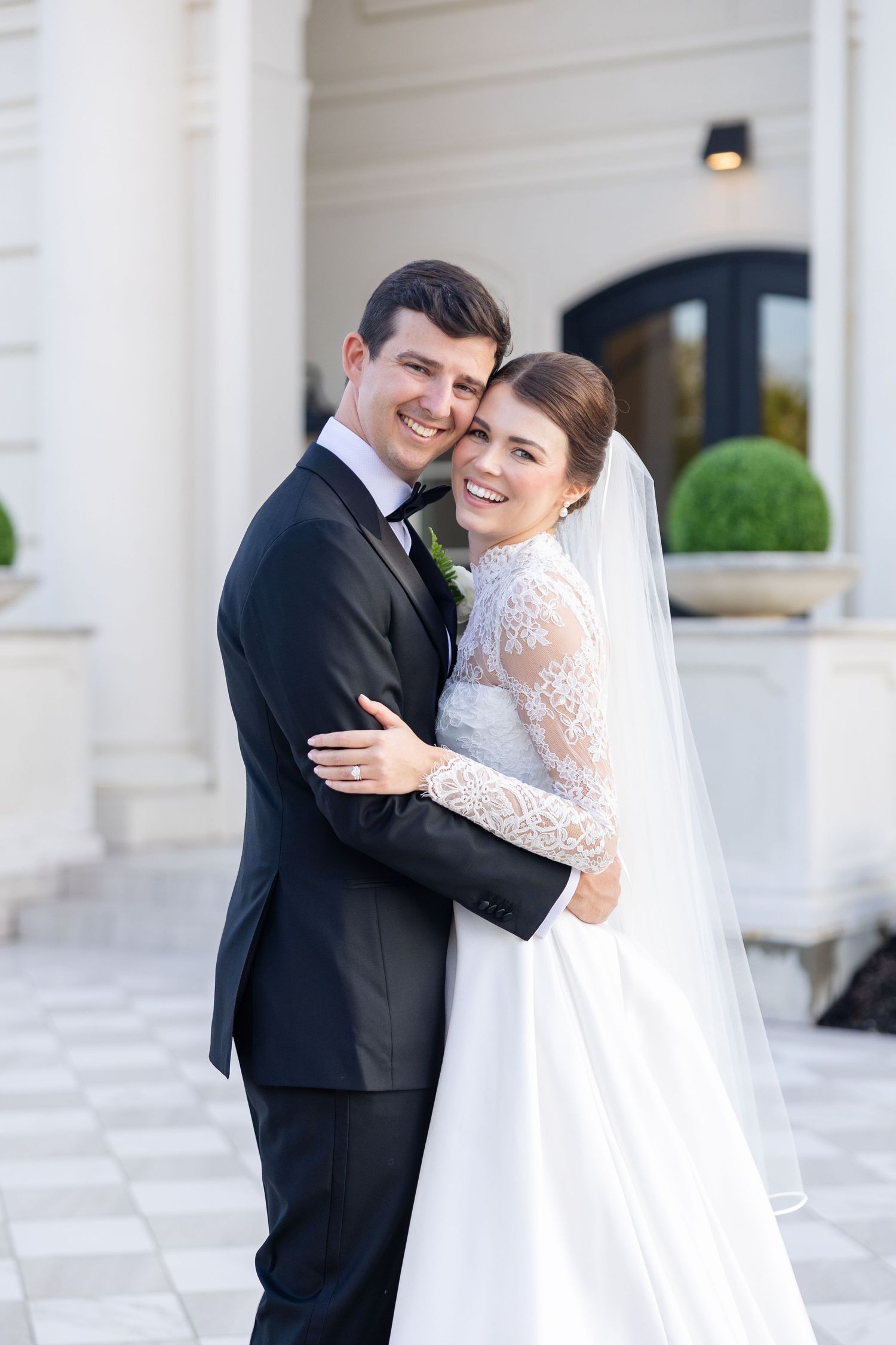 Happy bride and groom embracing and smiling at camera outside a classic white wedding venue.