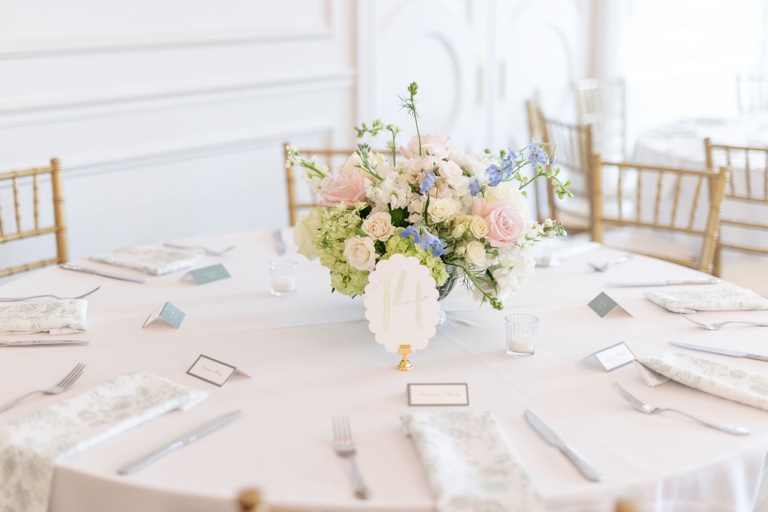 Elegant wedding reception table with blush pink and white floral centerpiece, gold chiavari chairs, and white linens.