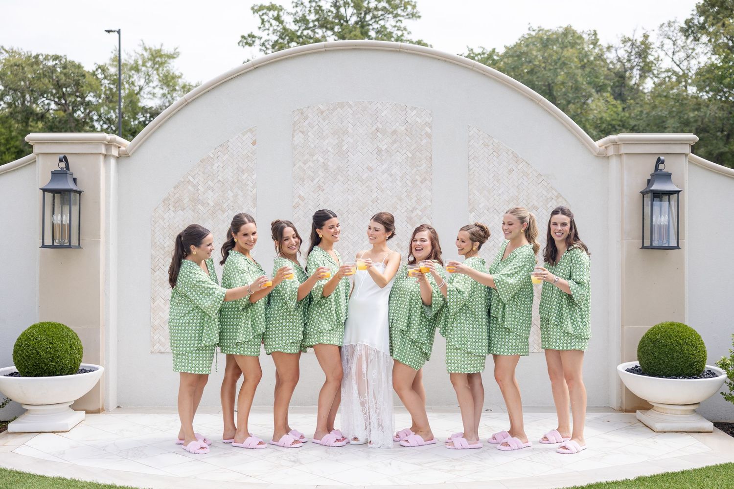 Bride in white stands with bridesmaids wearing matching green robes and pink slippers before an ornate arch.