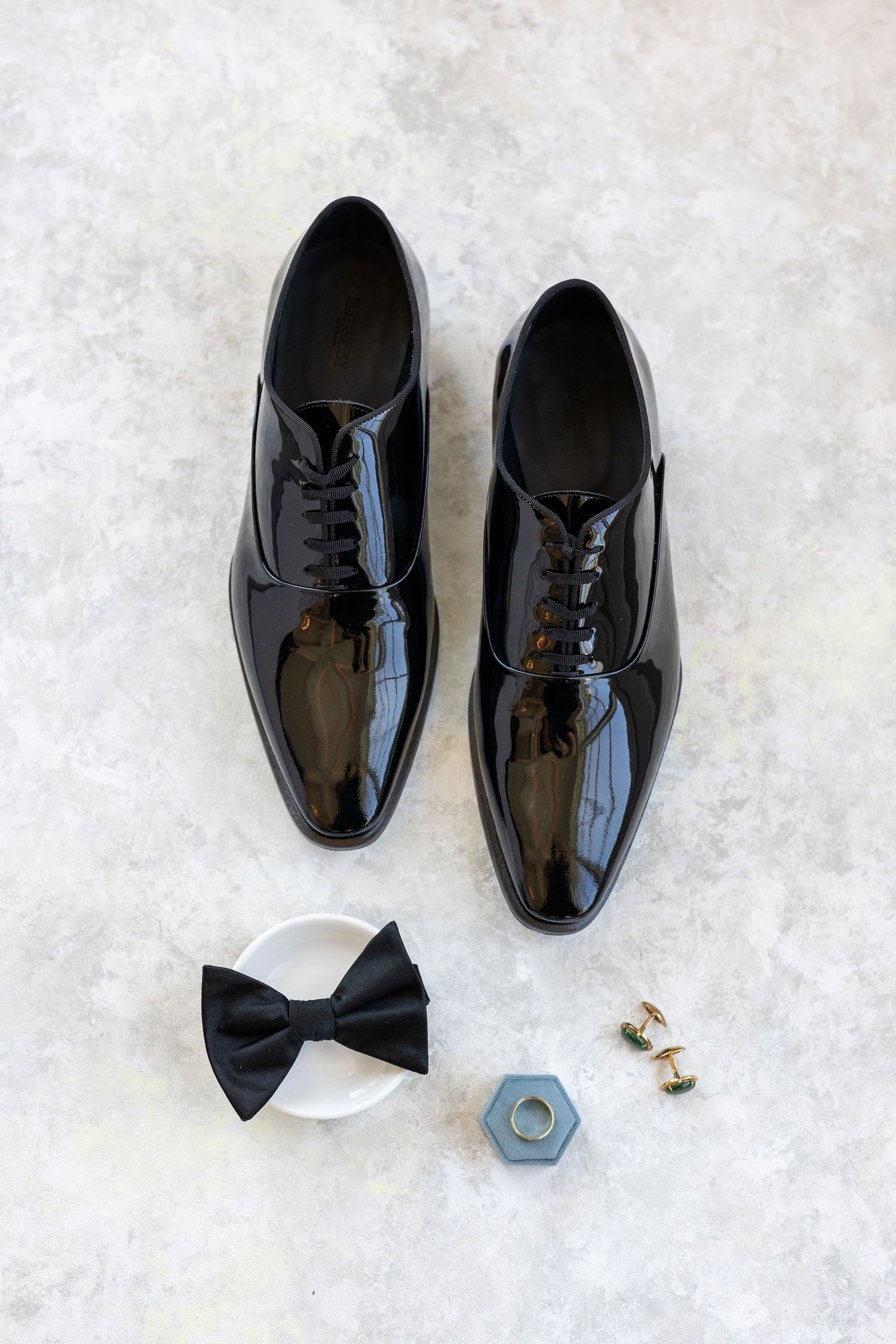 Black patent leather Oxford dress shoes with black bow tie and gold cufflinks on marble surface.
