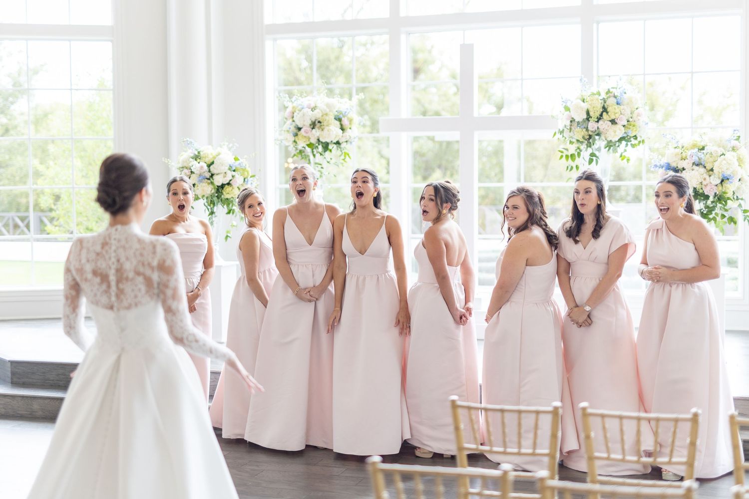 Bridesmaids in blush pink gowns reacting with joy upon seeing bride in white lace wedding dress.