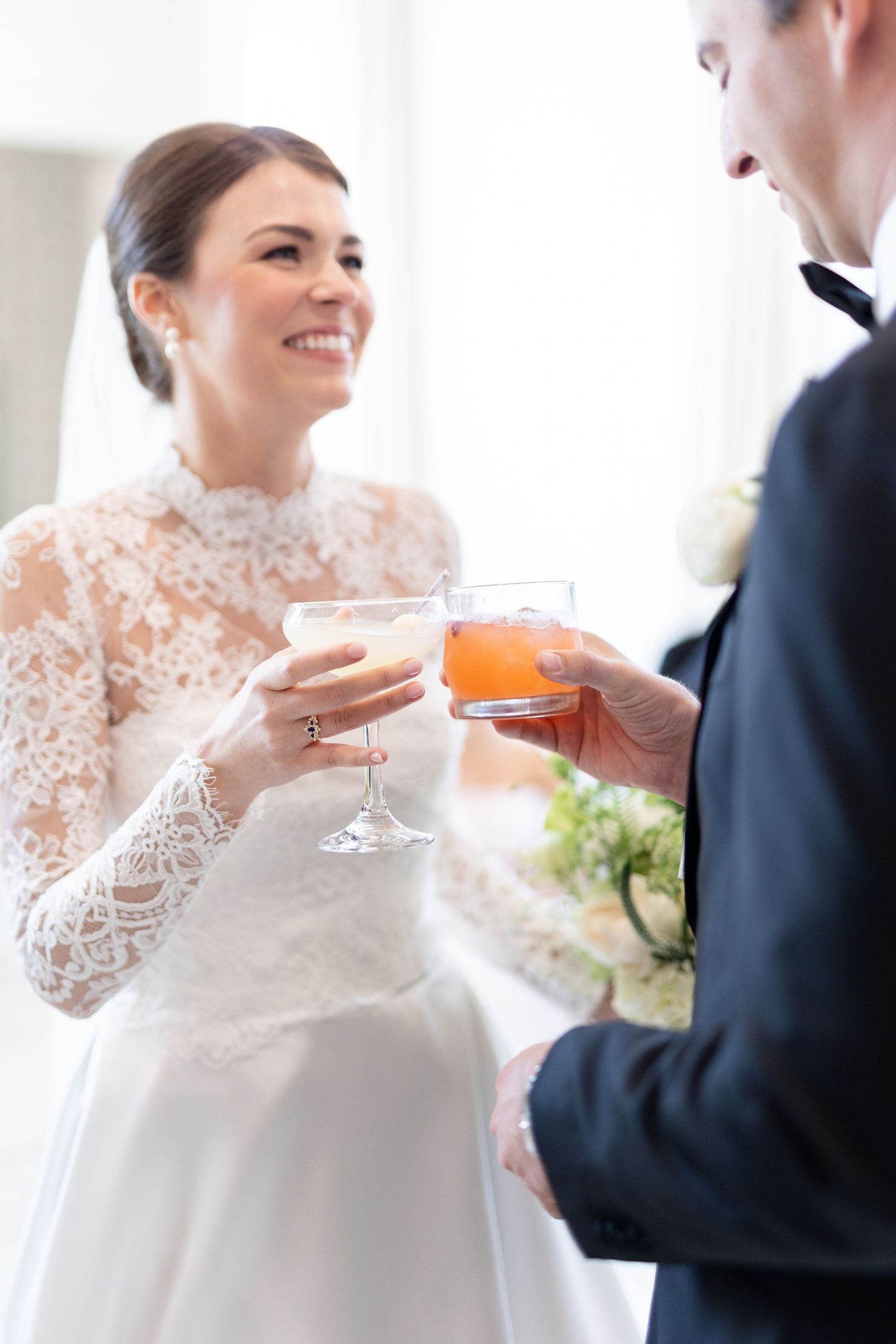 Smiling bride in lace wedding dress toasting with groom holding colorful cocktail glasses at wedding reception.