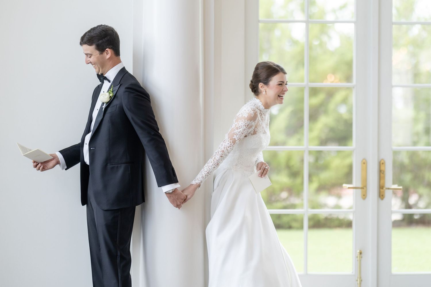 Bride and groom holding hands around white wall, groom reading vows before first look reveal.