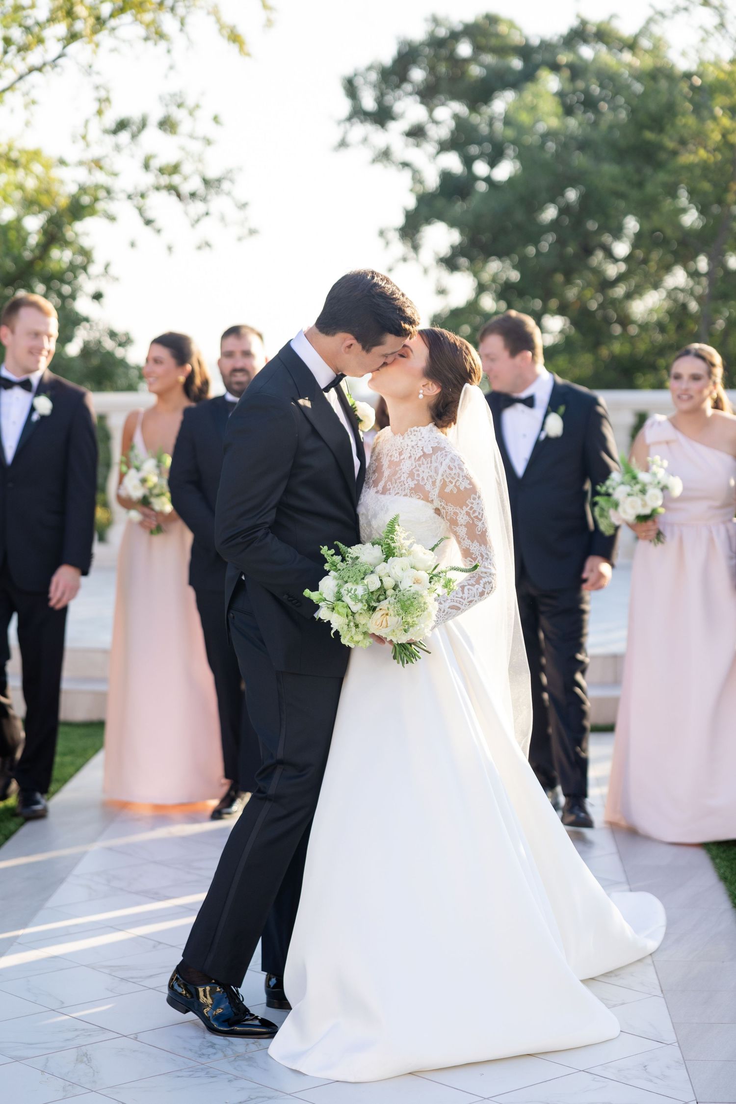 Bride and groom sharing a romantic kiss outdoors with wedding party in blush and black attire in background.