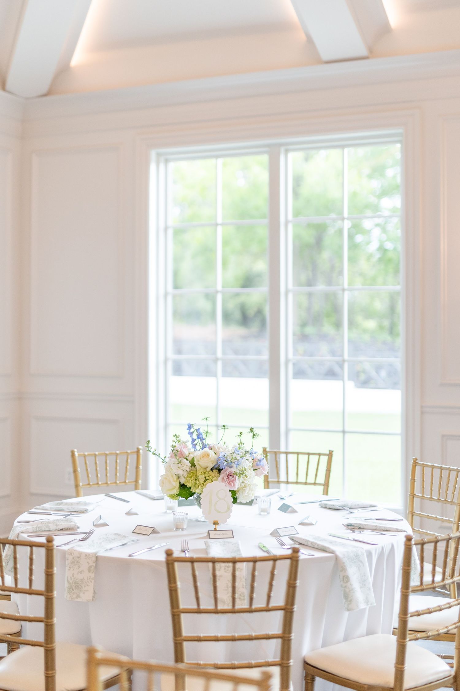 Bright white wedding reception room with gold chiavari chairs, round table, and pastel floral centerpiece by window.