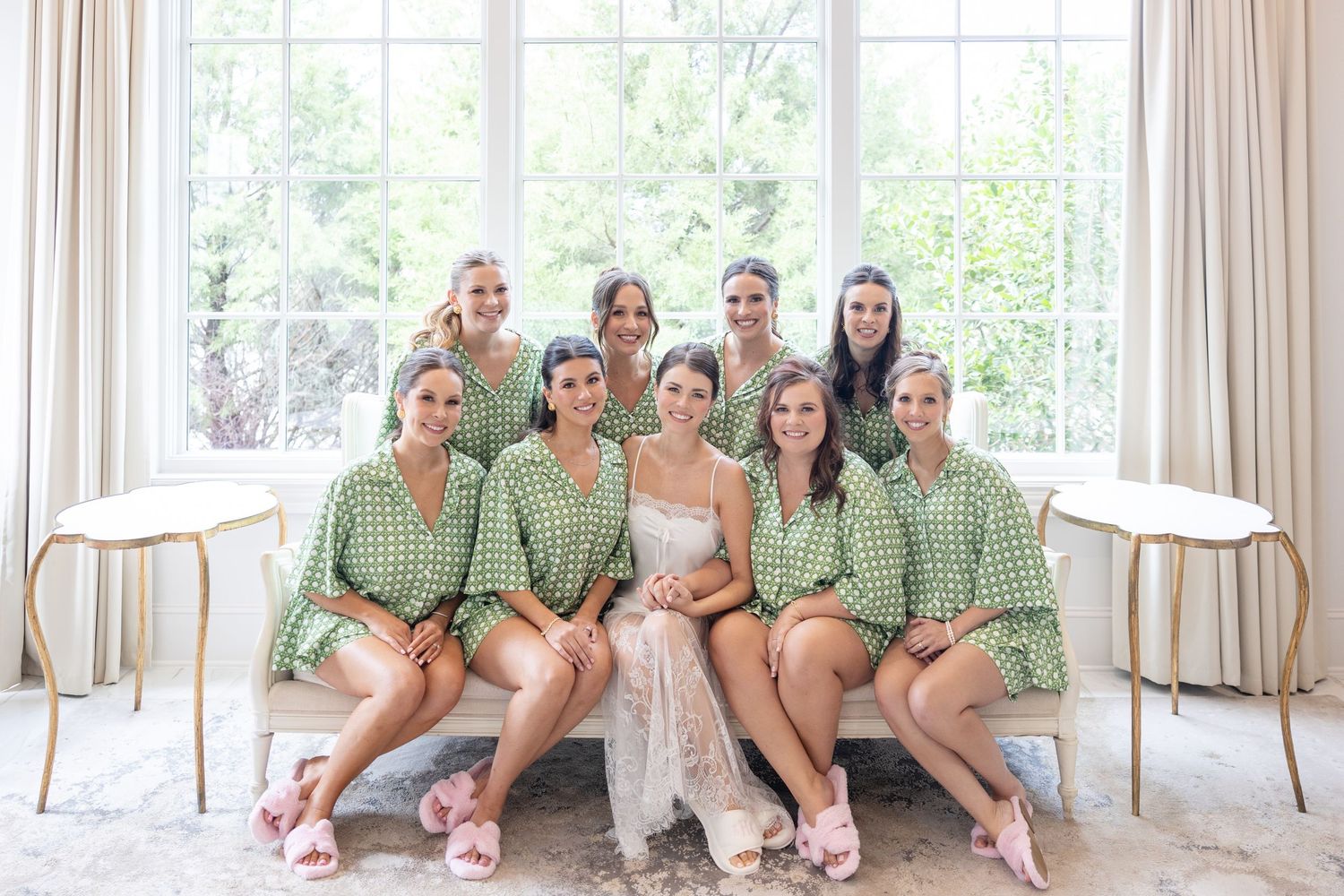 Bride poses with nine bridesmaids in matching green robes seated and standing in a bright elegant room.