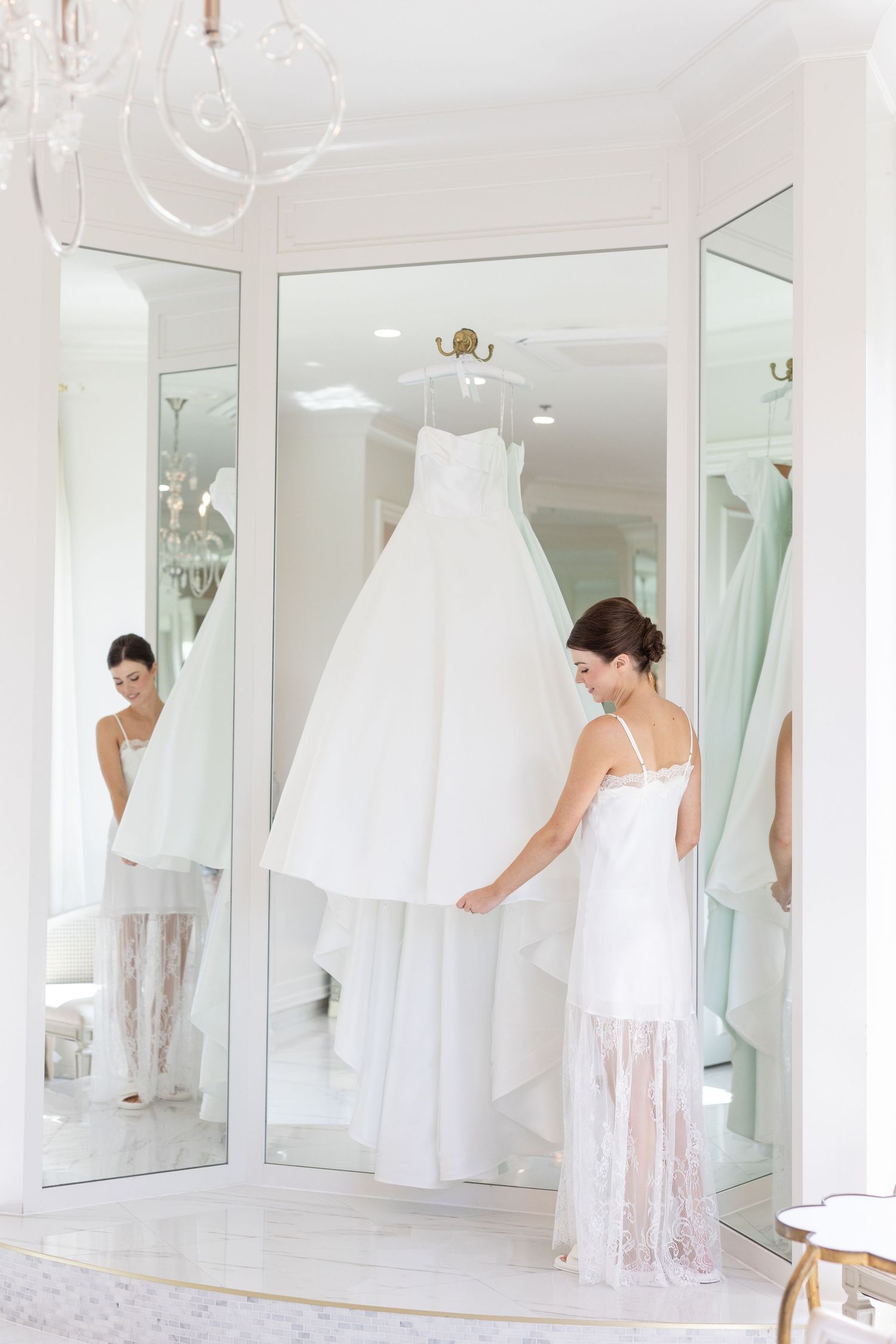 Bride in slip dress admiring her white ball gown wedding dress hanging in mirrored dressing room.