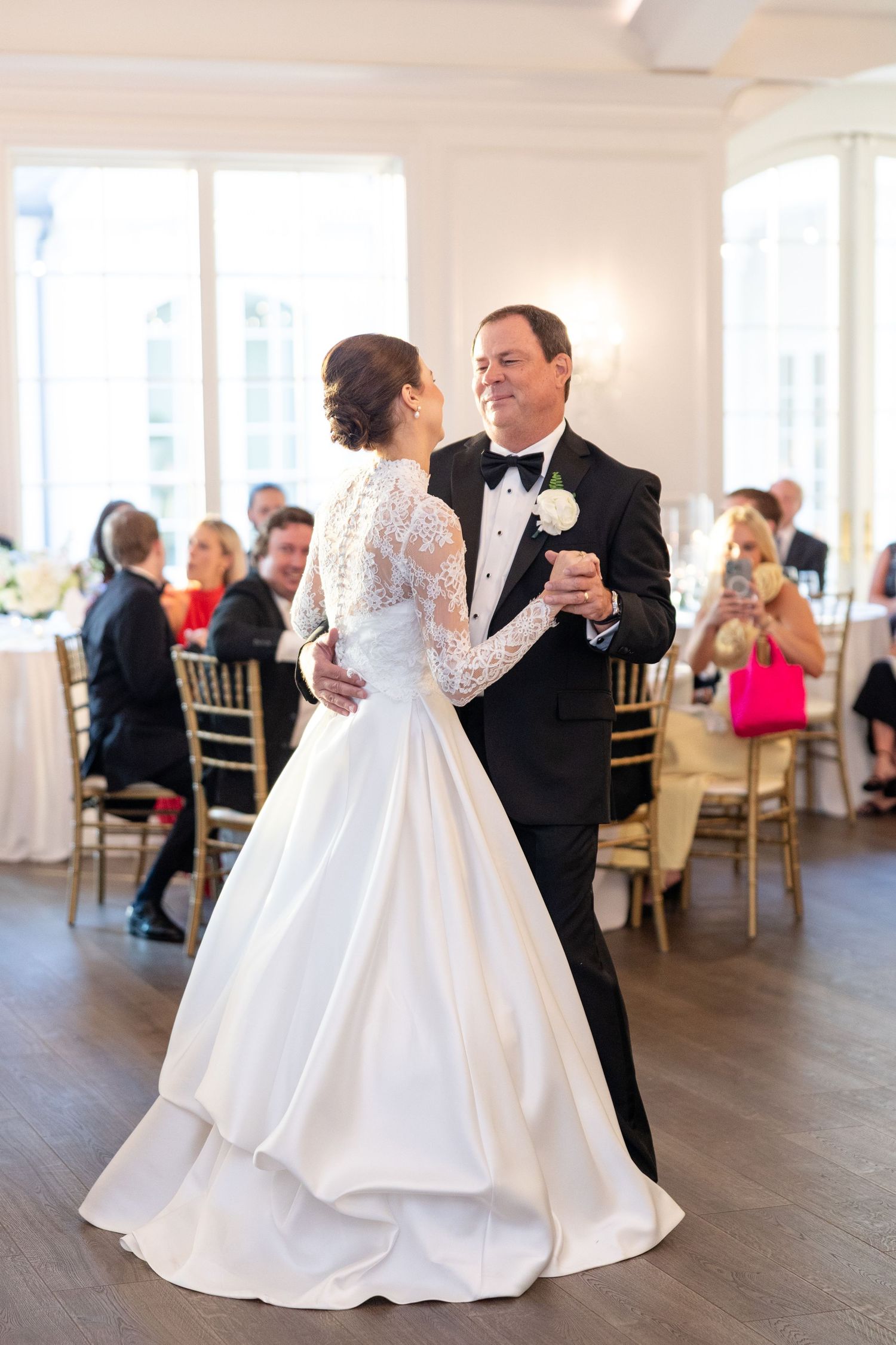 Bride in long-sleeve lace gown shares father-daughter dance in elegant white ballroom with seated wedding guests.