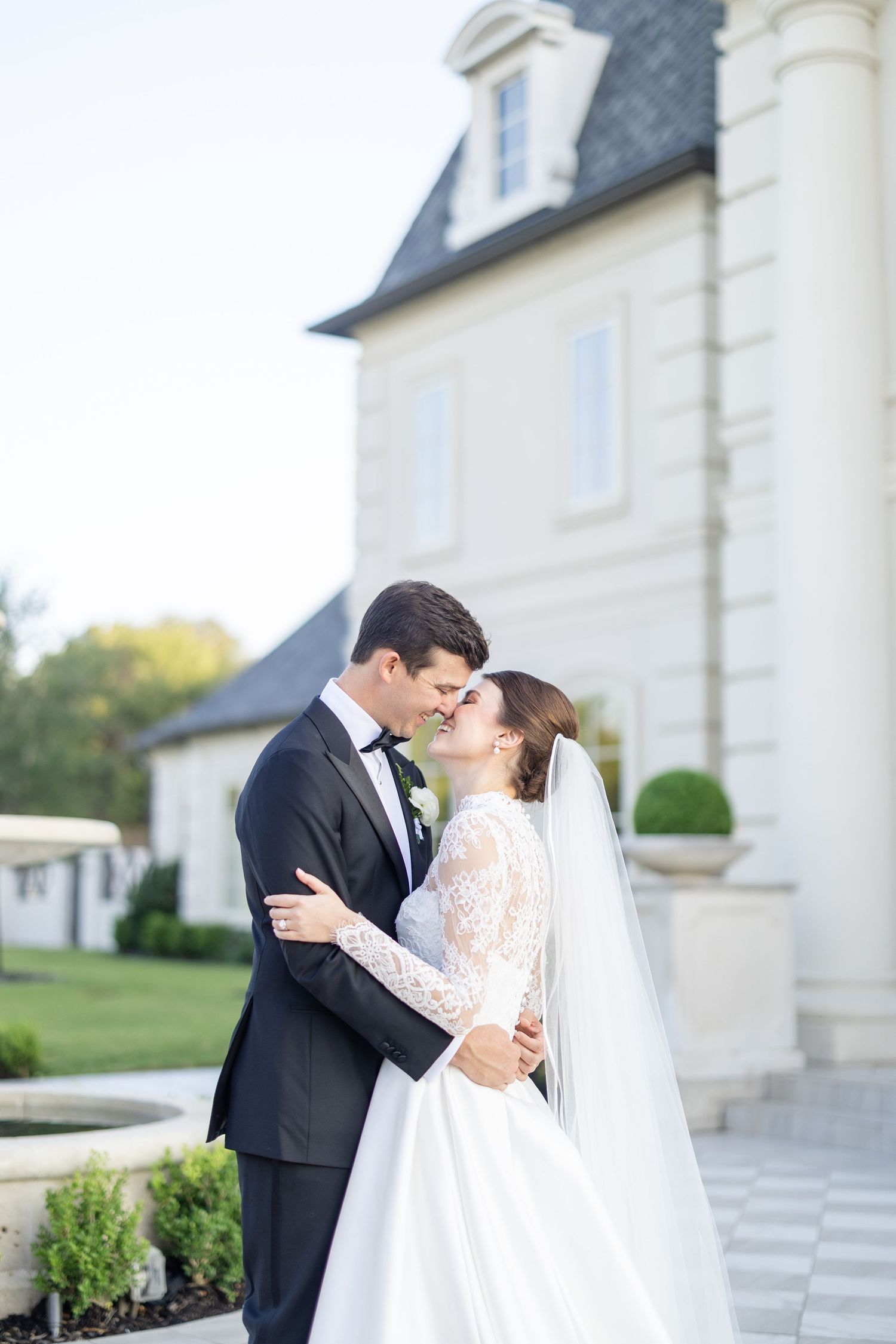 Bride in lace long-sleeve gown and groom in tuxedo sharing an intimate moment outside a chateau.