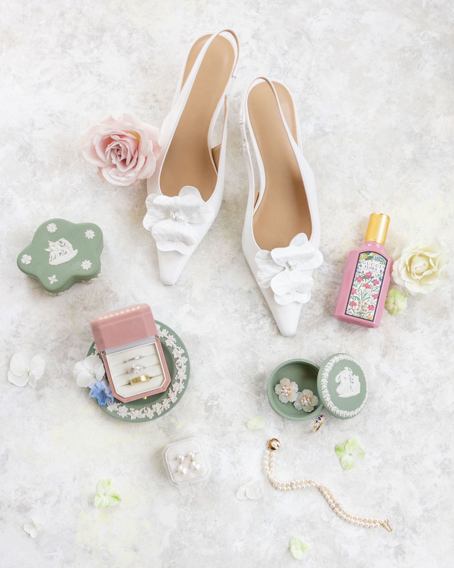 Bridal flat lay featuring white bow heels, diamond ring, pearl bracelet, green trinket boxes, and pink perfume.