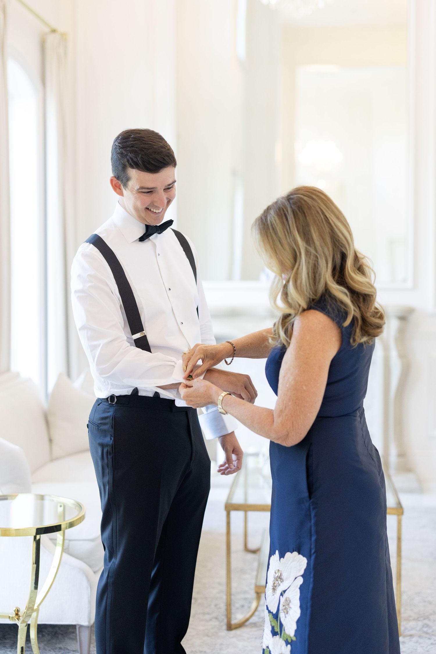 Groom in suspenders and bow tie smiles as woman in navy dress adjusts his cufflinks before wedding.