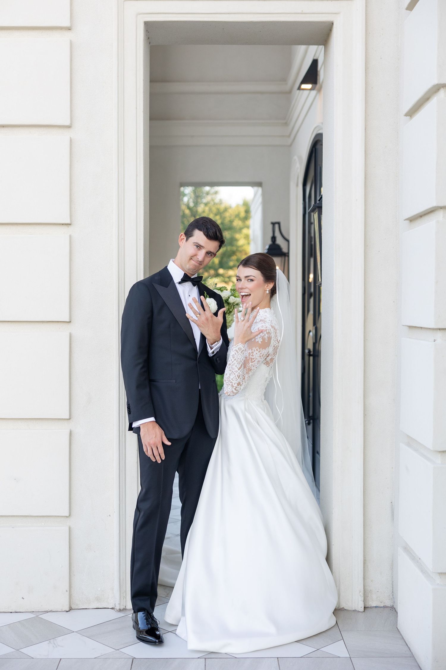 Bride in lace ballgown and groom in black tuxedo posing playfully in a white architectural doorway.
