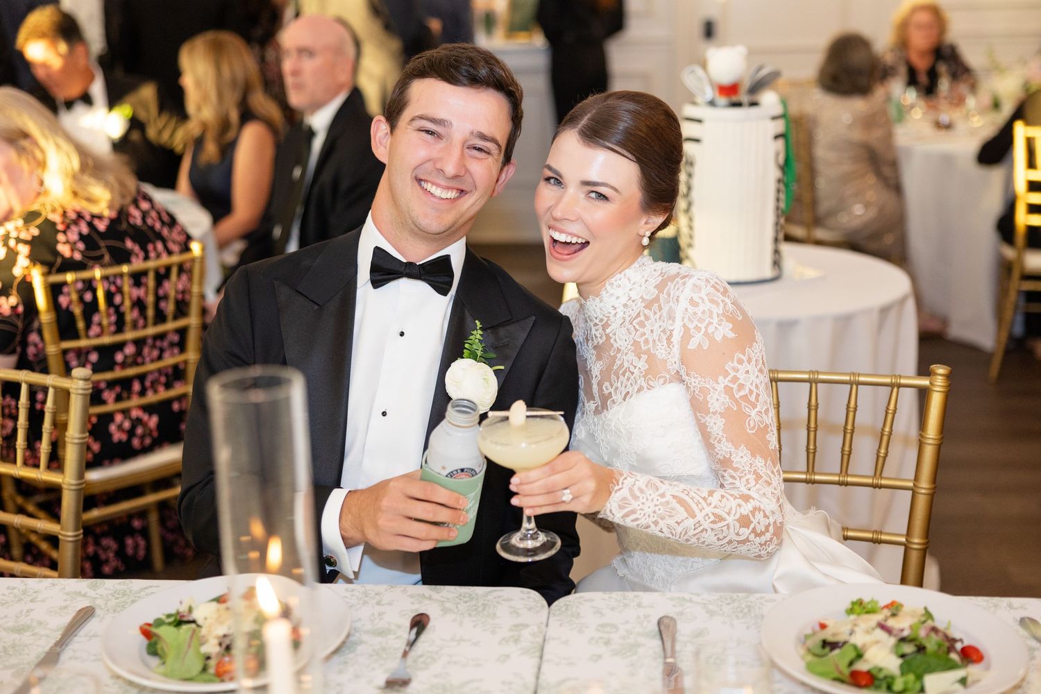 Smiling bride and groom toast with cocktails while seated at their sweetheart table during wedding reception.