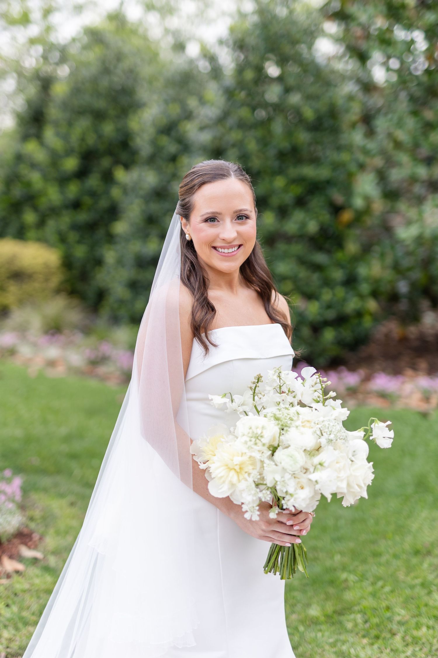 Elegant bride in strapless white gown holding a lush white floral bouquet in a garden setting.