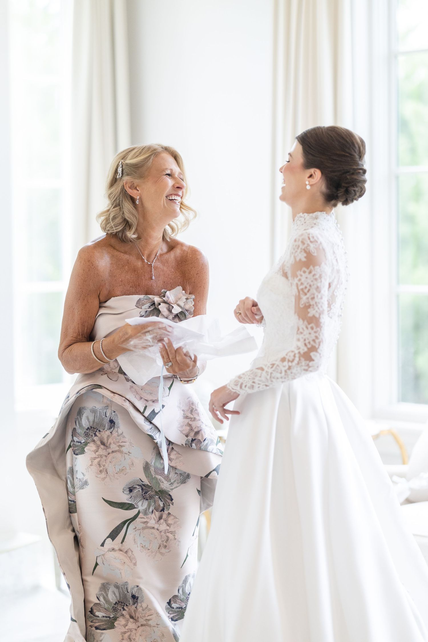 Bride in lace wedding gown laughing with mother in floral dress during bridal preparations.