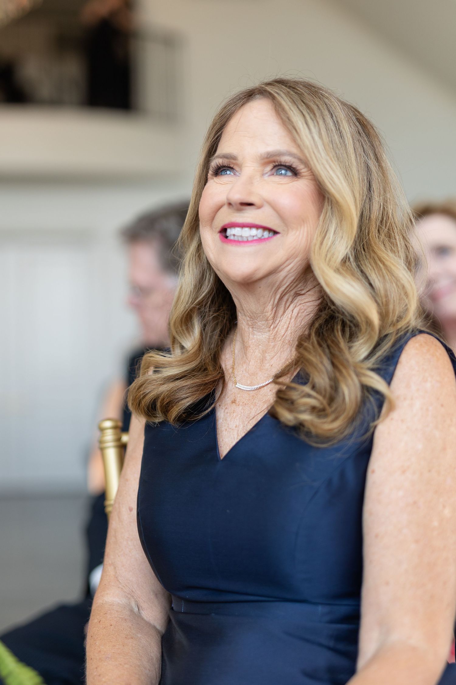Smiling blonde woman in navy dress enjoying a wedding ceremony seated among other guests.