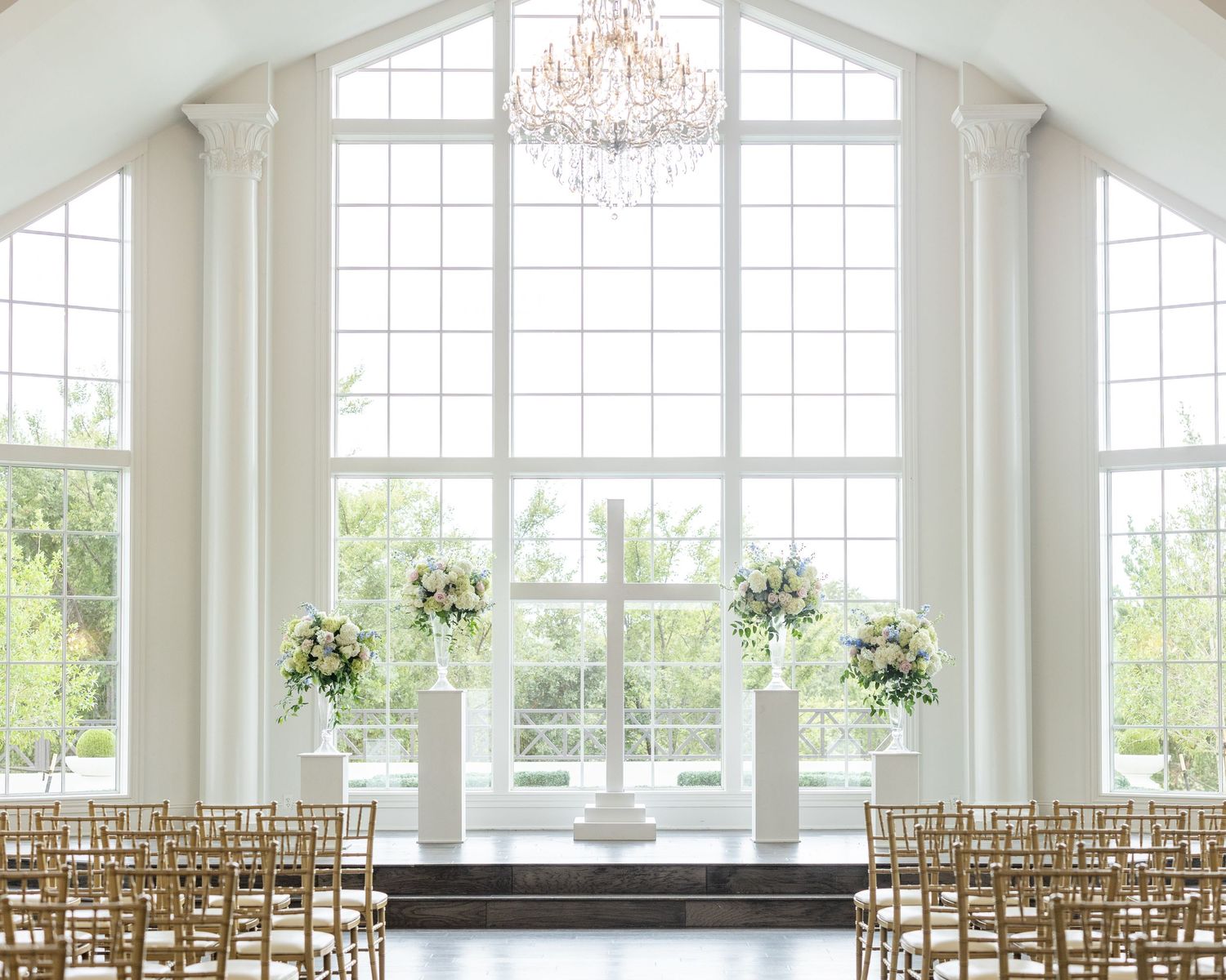 All-white wedding ceremony chapel with crystal chandelier, tall windows, and gold chiavari chairs.