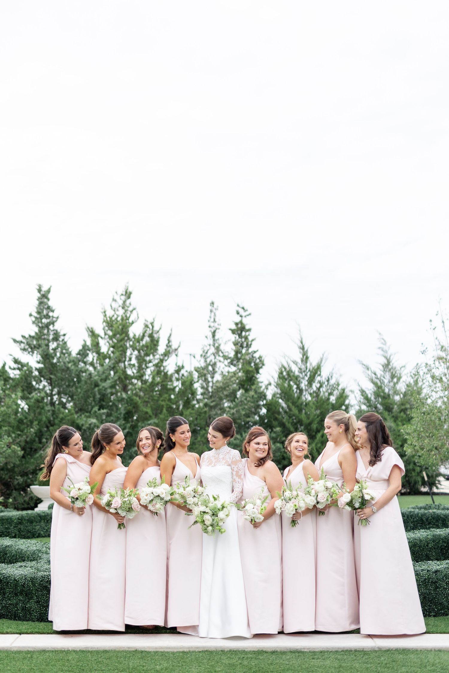 Bride in lace wedding gown surrounded by bridesmaids in blush dresses holding white bouquets.