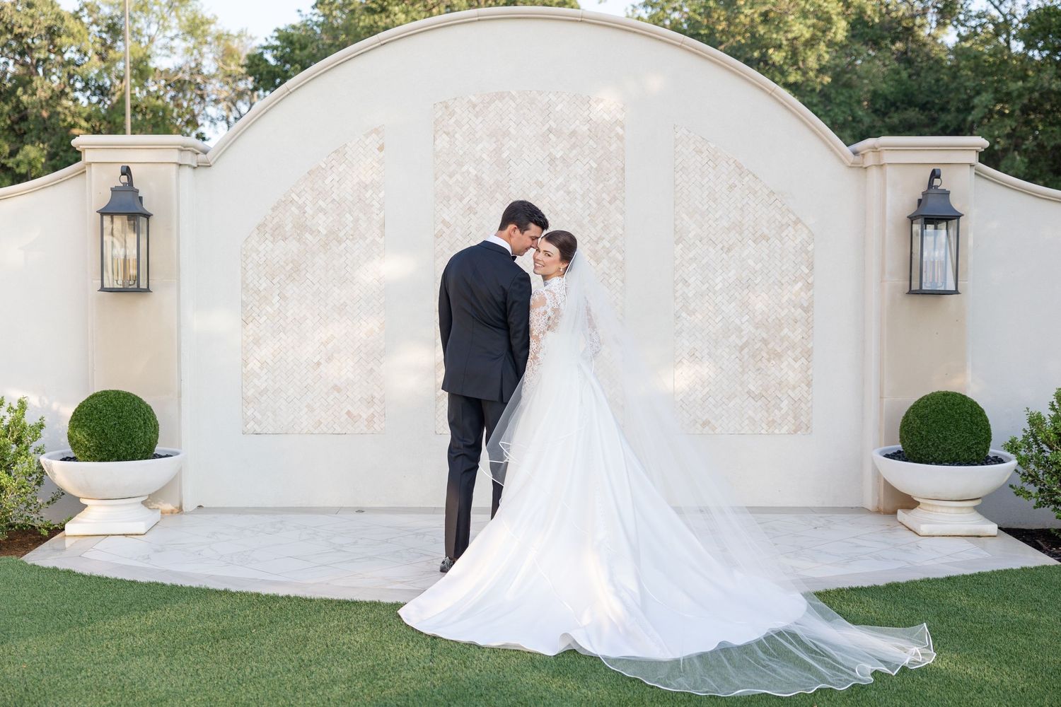 Bride and groom embrace tenderly before a grand white arched wall with lanterns at sunset.