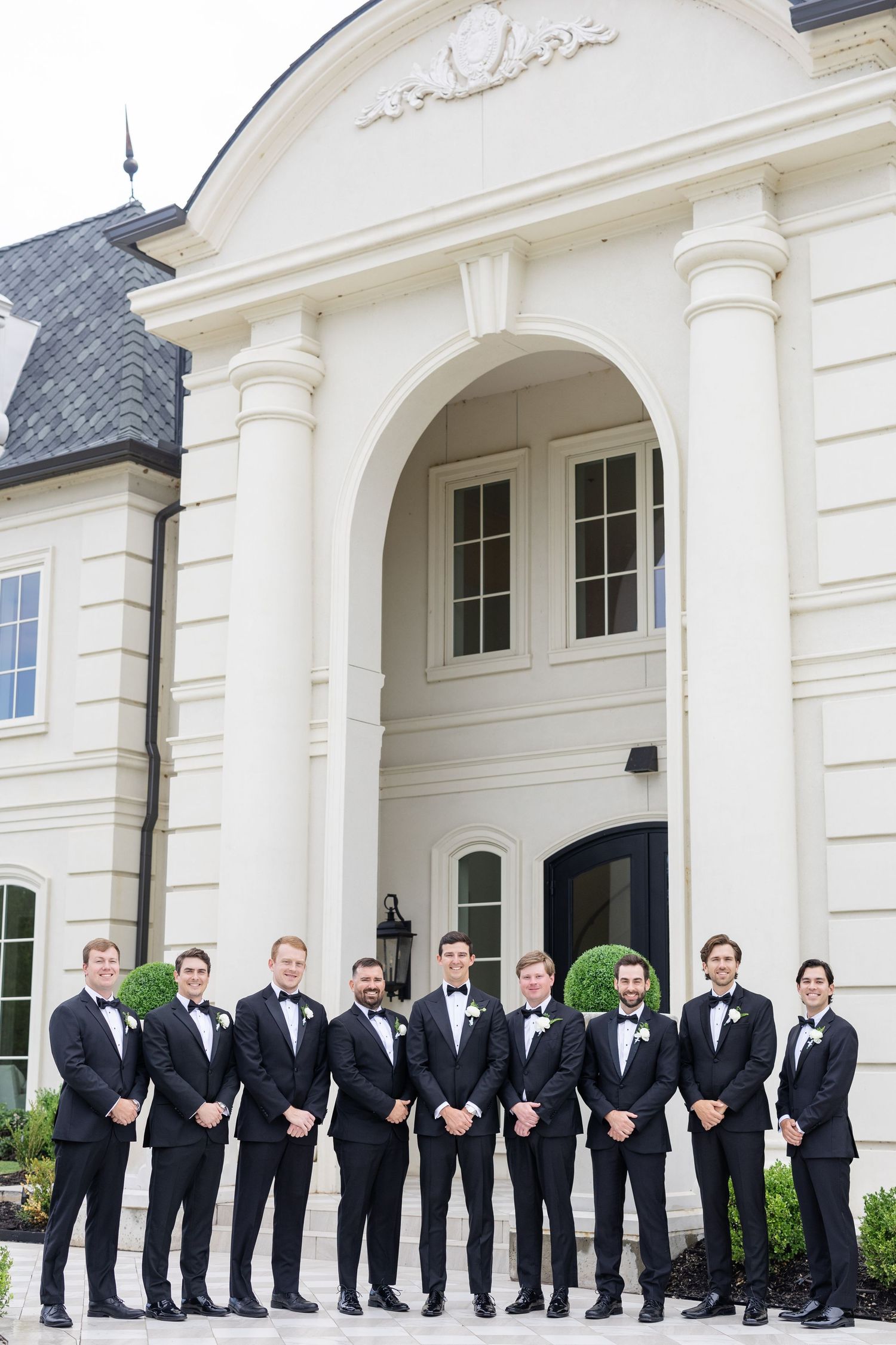 Groomsmen in black tuxedos with bow ties pose in front of an elegant white mansion entrance.
