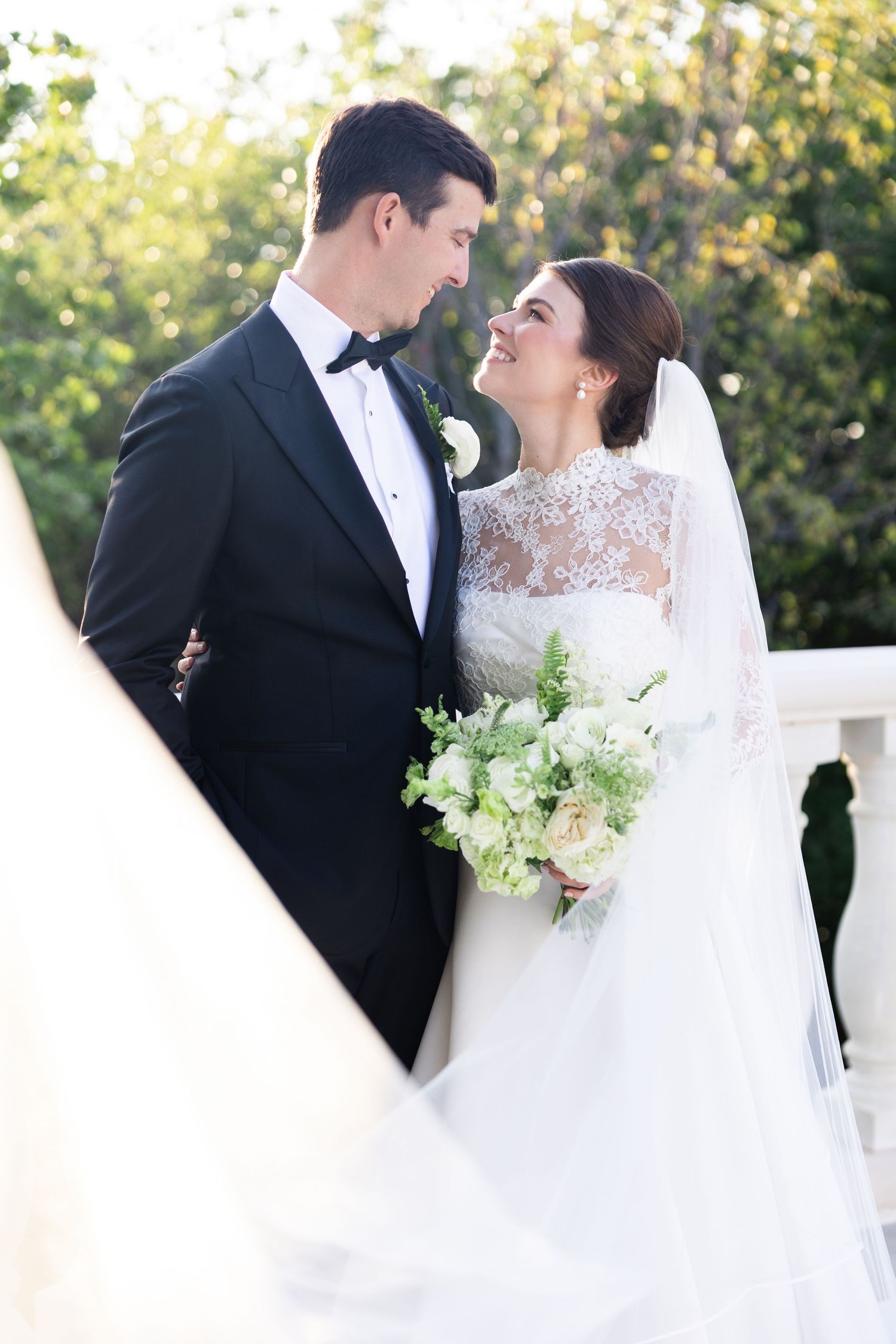 Smiling bride in lace wedding gown and groom in tuxedo share a tender moment on a balcony.
