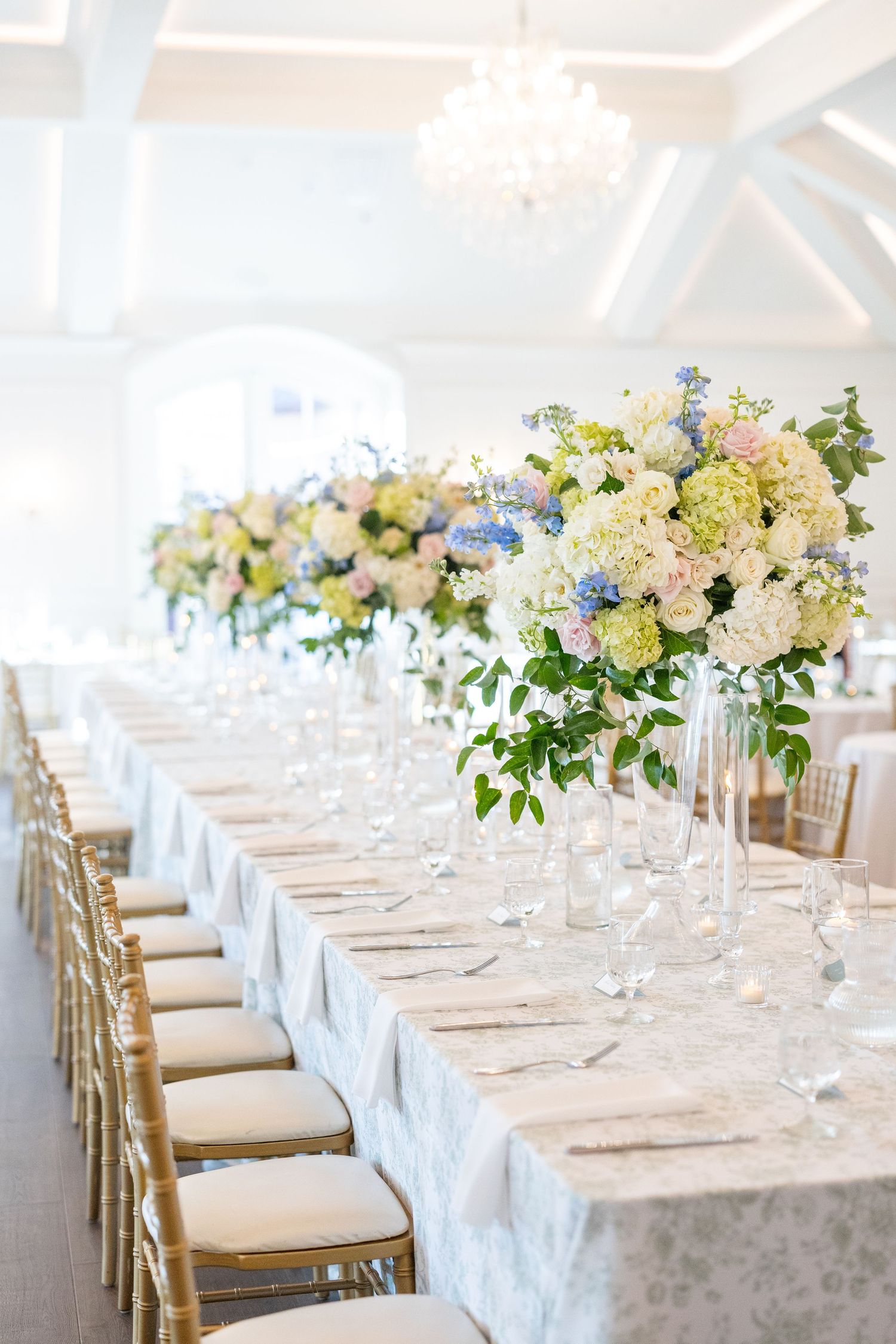 Long reception table with tall floral centerpieces of white, pink, and blue blooms and gold chiavari chairs.