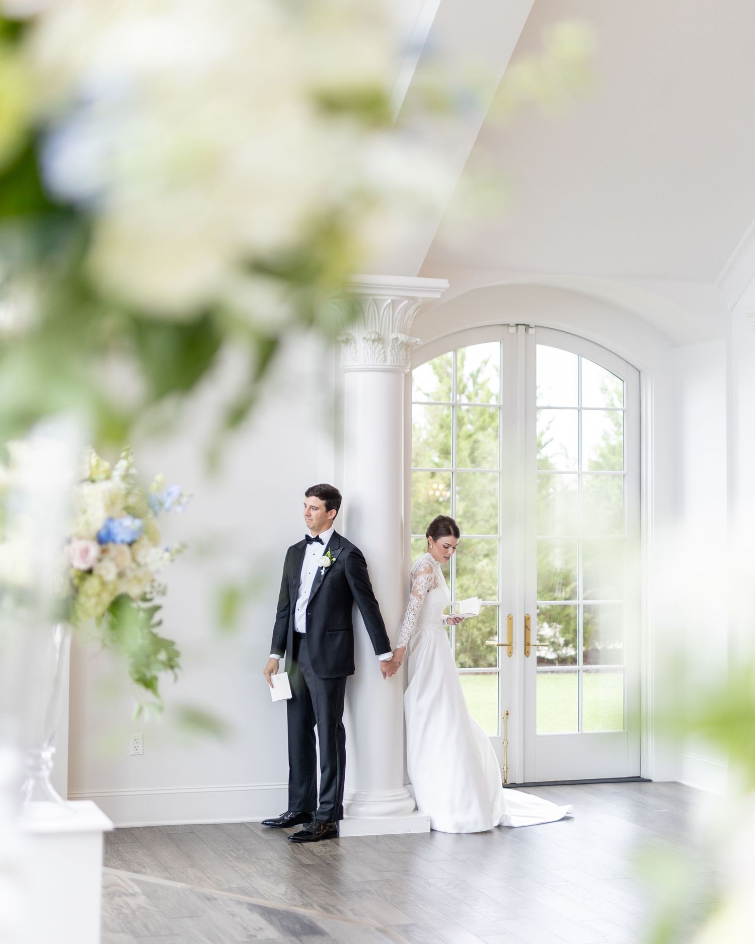 Bride in lace gown and groom in black tuxedo holding hands near arched French doors with floral arrangements.