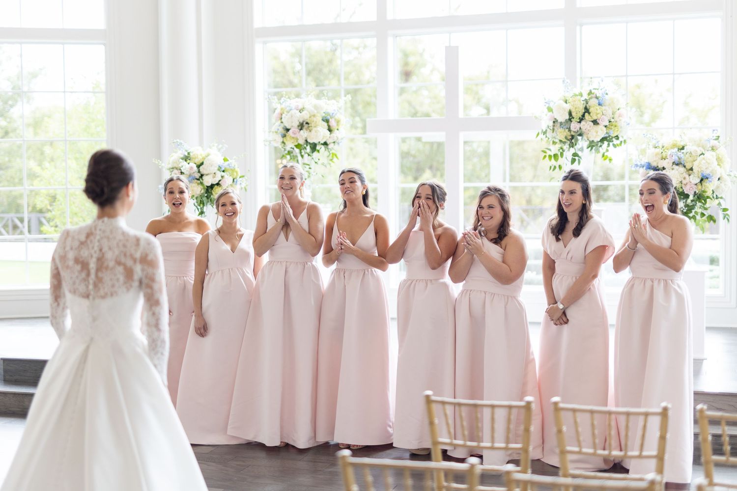 Seven bridesmaids in blush pink dresses clapping with excitement at bride in lace wedding gown.