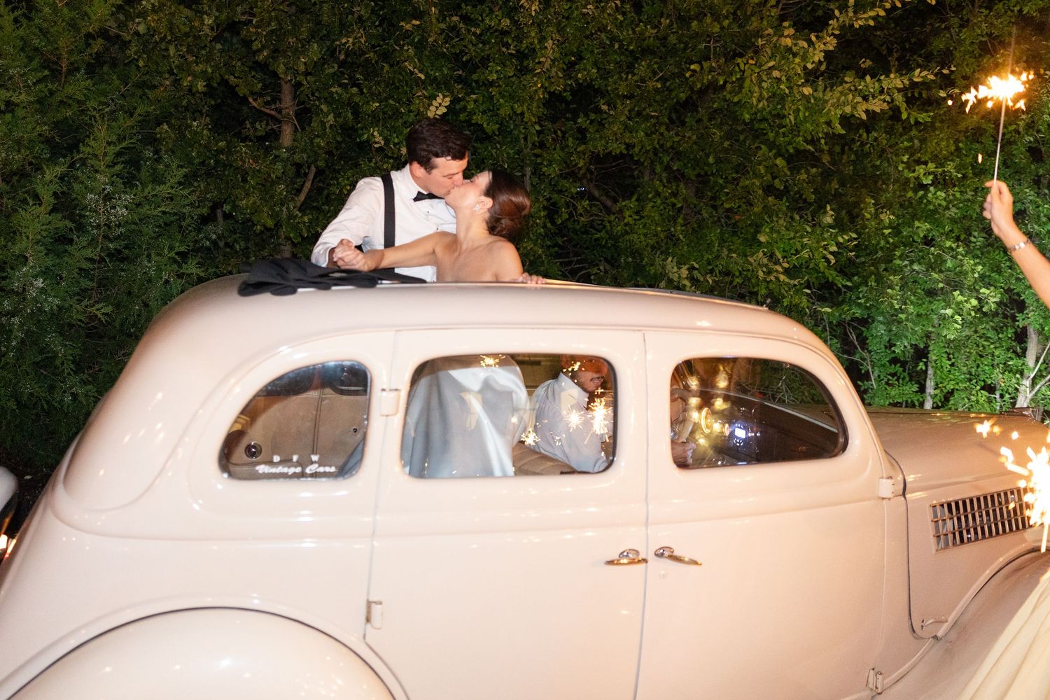 Newlyweds kiss through sunroof of vintage cream getaway car surrounded by sparklers.