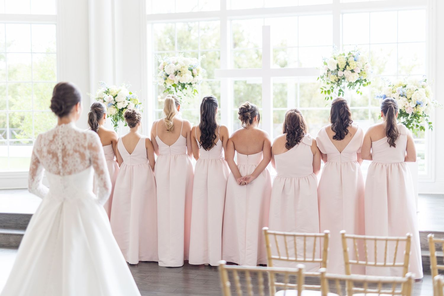 Bride facing seven bridesmaids in blush pink gowns standing before bright windows with florals.