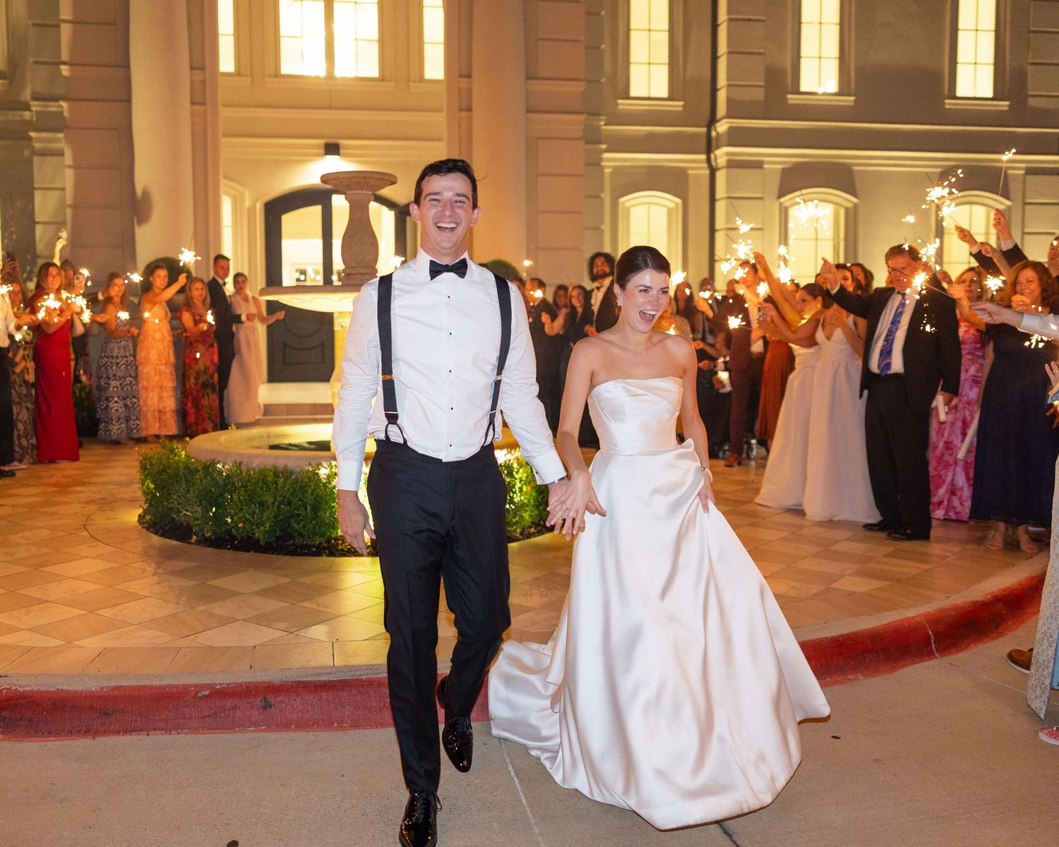 Bride and groom exit wedding venue through sparkler send-off tunnel with cheering guests.