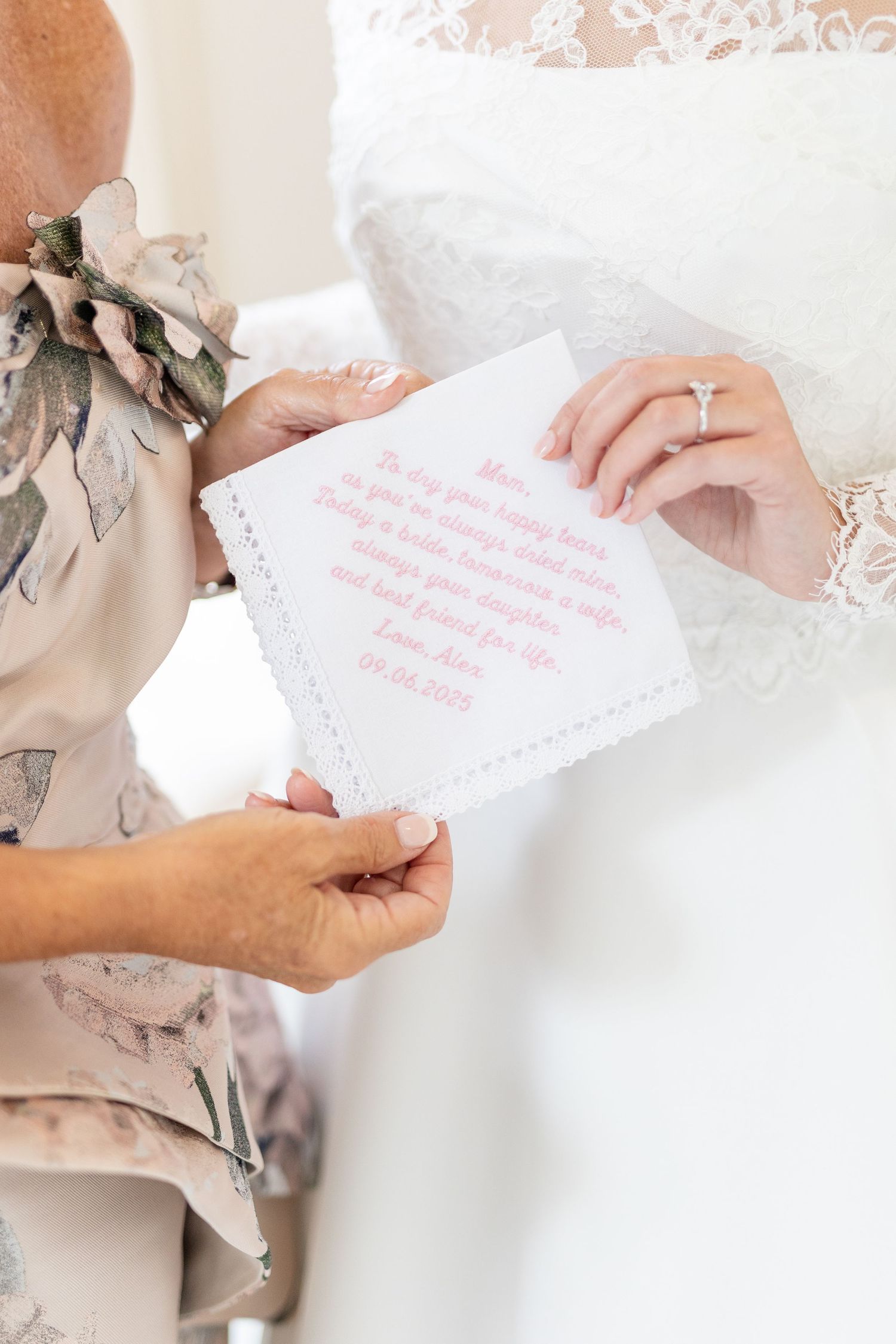 Bride and mother sharing embroidered handkerchief with pink cursive message on wedding day.