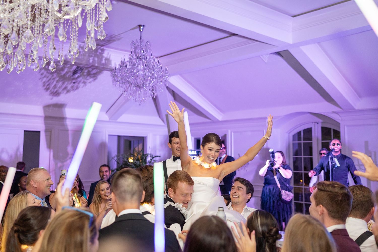 Bride raises arms joyfully on dance floor surrounded by guests with glow sticks at elegant wedding reception.