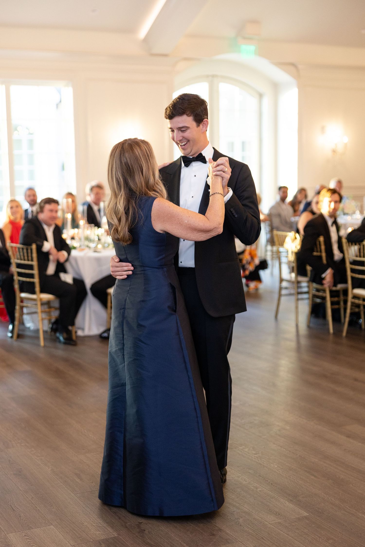 Groom in tuxedo shares a joyful mother-son dance in bright white wedding reception ballroom with guests watching.