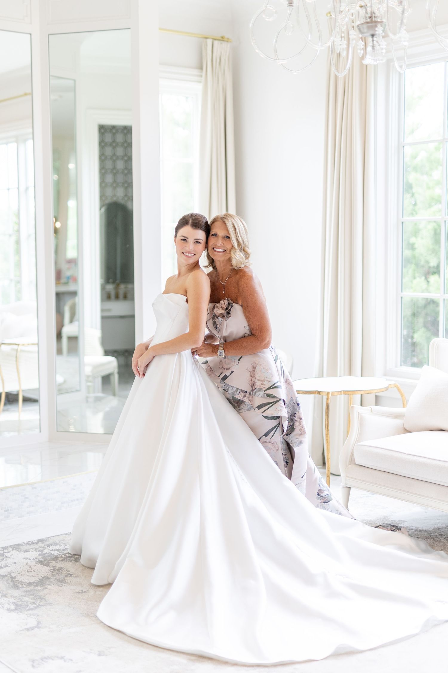 Bride in white strapless ball gown posing with smiling mother in floral dress in bright elegant room.