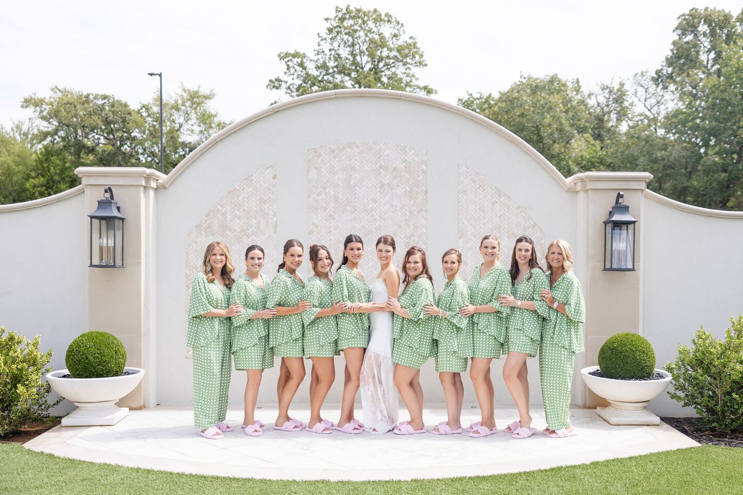 Bride and bridesmaids in green robes and pink slippers pose in a line before a decorative venue arch.