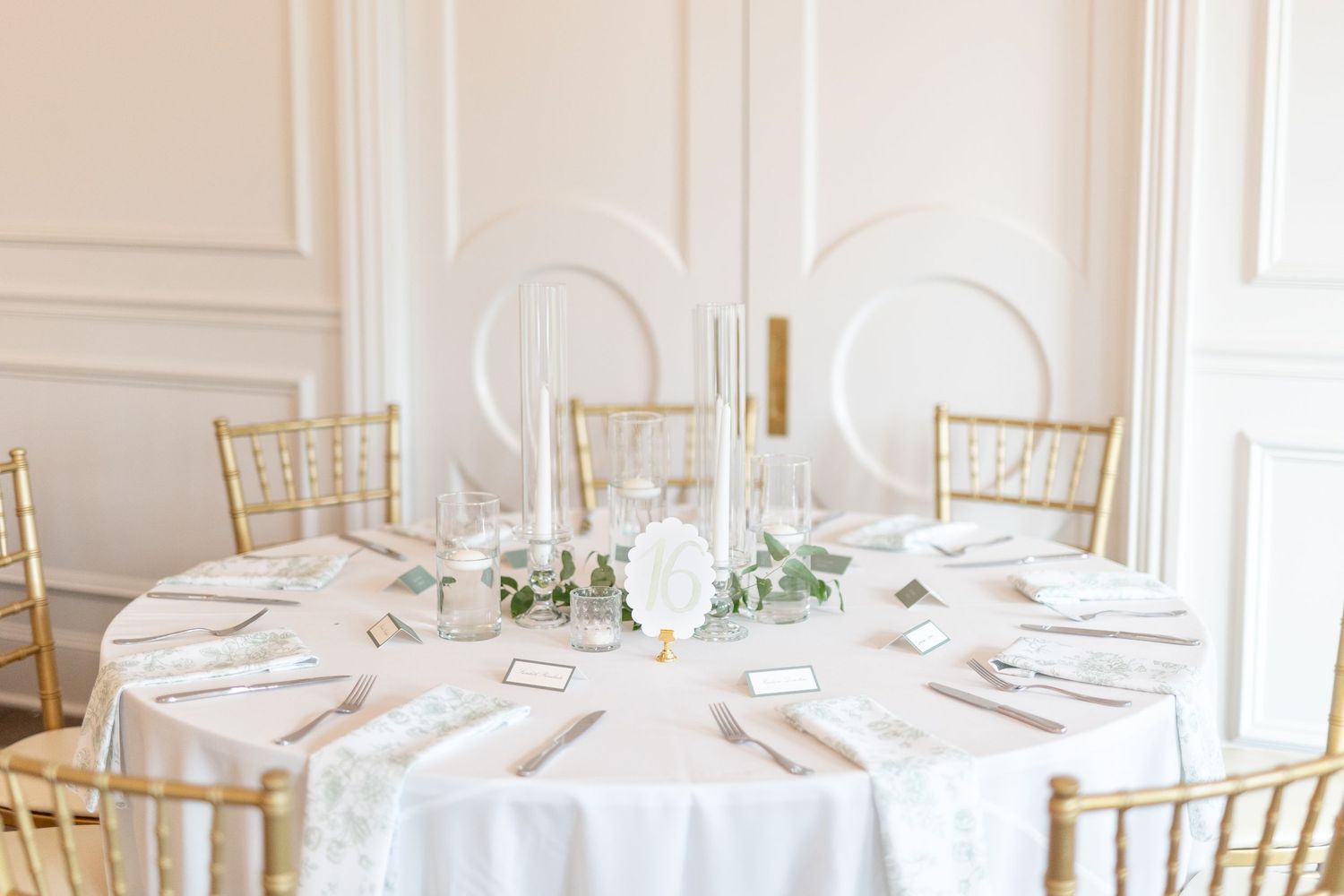 Classic white wedding reception table with tall taper candles, greenery centerpiece, and gold chiavari chairs.