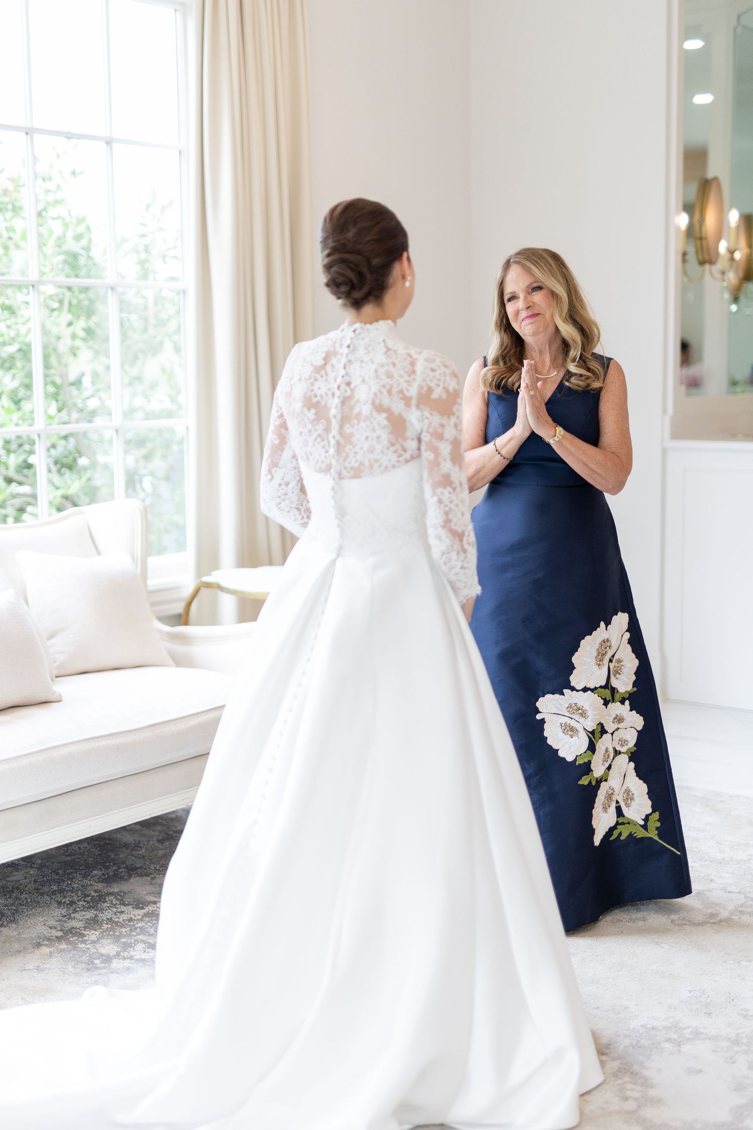Bride in lace long-sleeve gown standing before woman in navy floral dress with clasped hands.