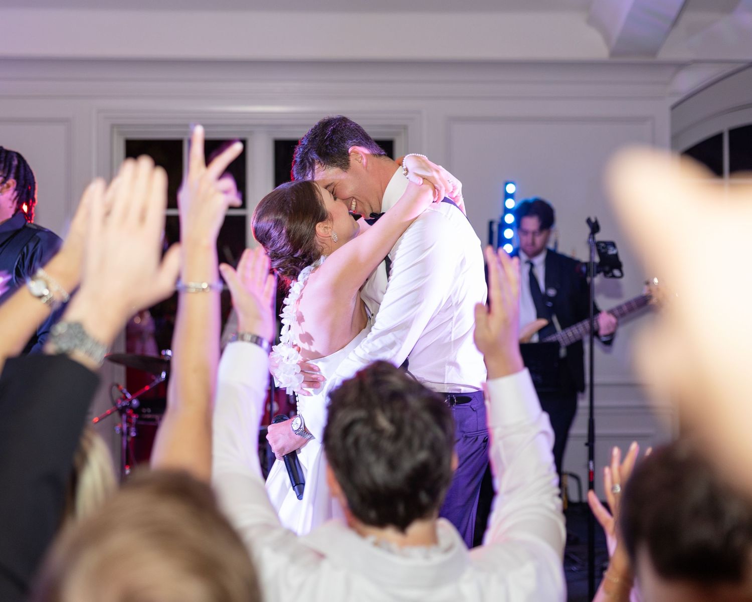 Bride and groom share romantic kiss on dance floor surrounded by cheering wedding guests.