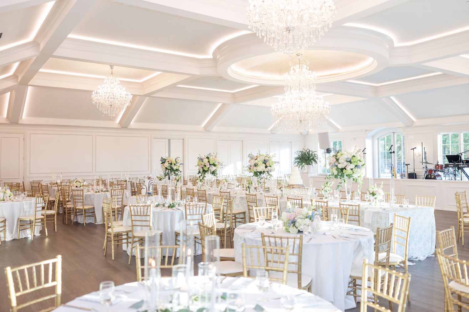 Elegant wedding reception ballroom with gold chiavari chairs, white round tables, crystal chandeliers, and tall floral centerpieces.