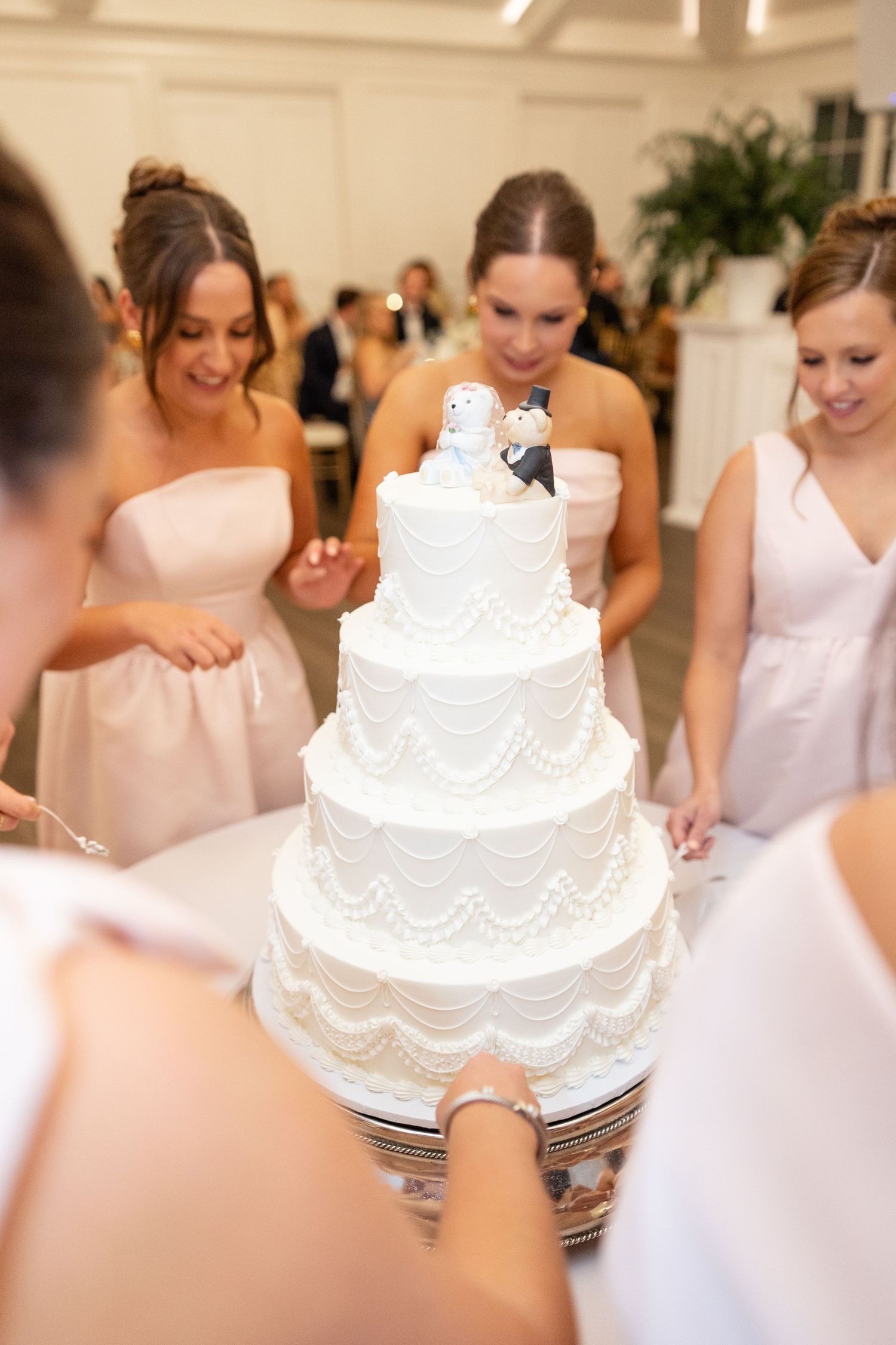 Bridesmaids in blush gowns admiring four-tier white wedding cake with bride and groom topper.