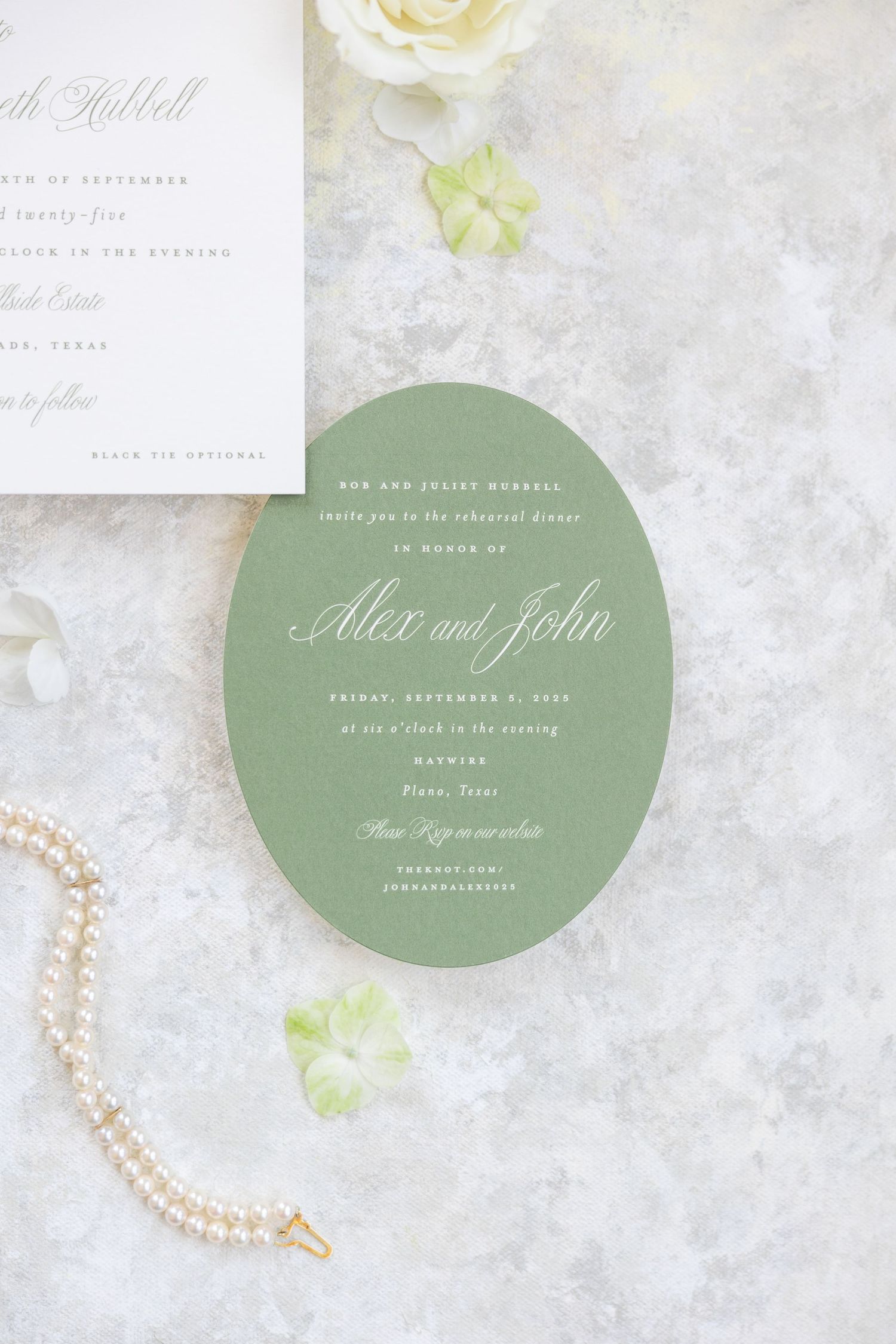 Sage green oval rehearsal dinner invitation for Alex and John displayed with pearl necklace and white roses.