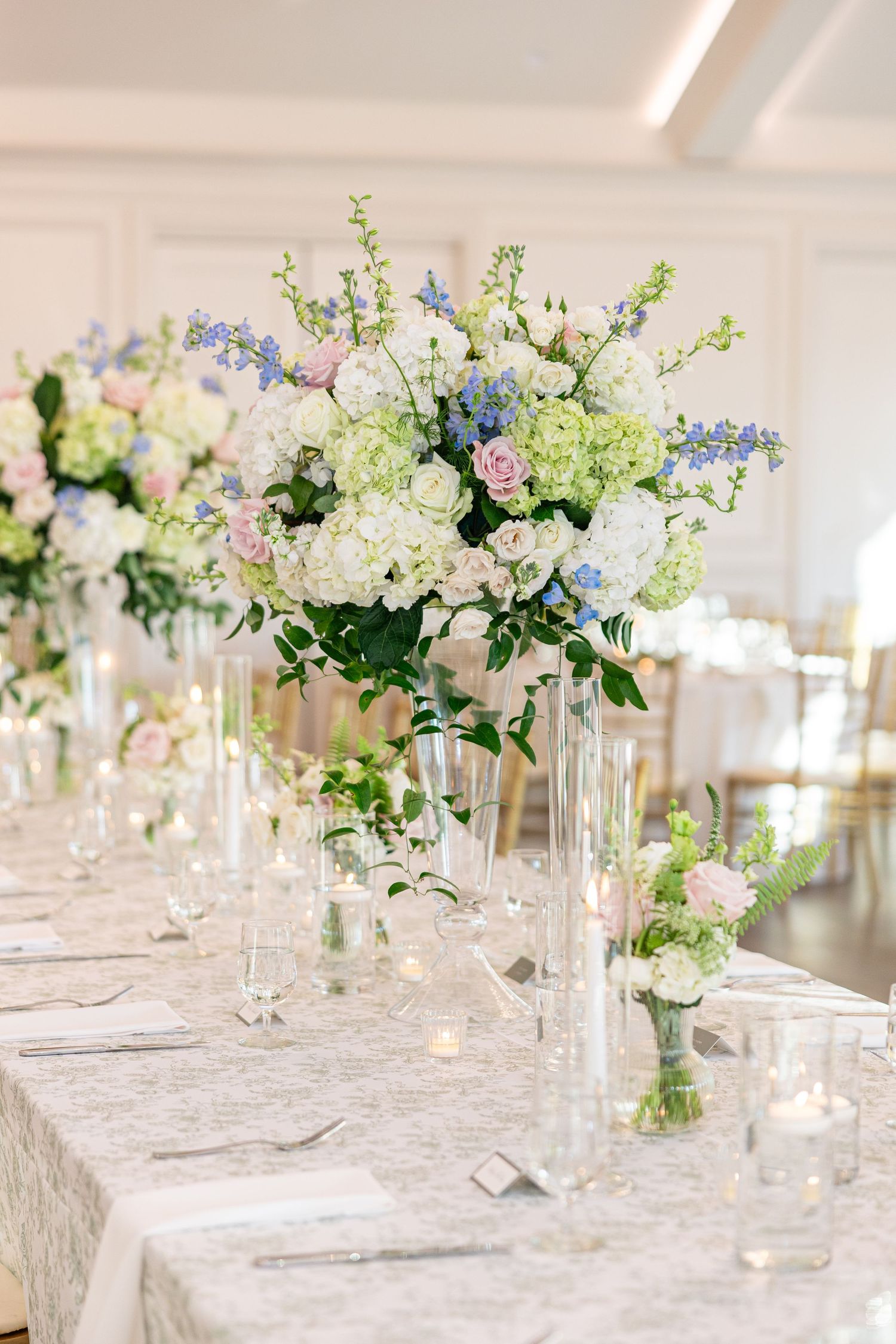 Tall glass vase centerpiece with white hydrangeas, blue delphinium, and pink roses on a lace-covered reception table.
