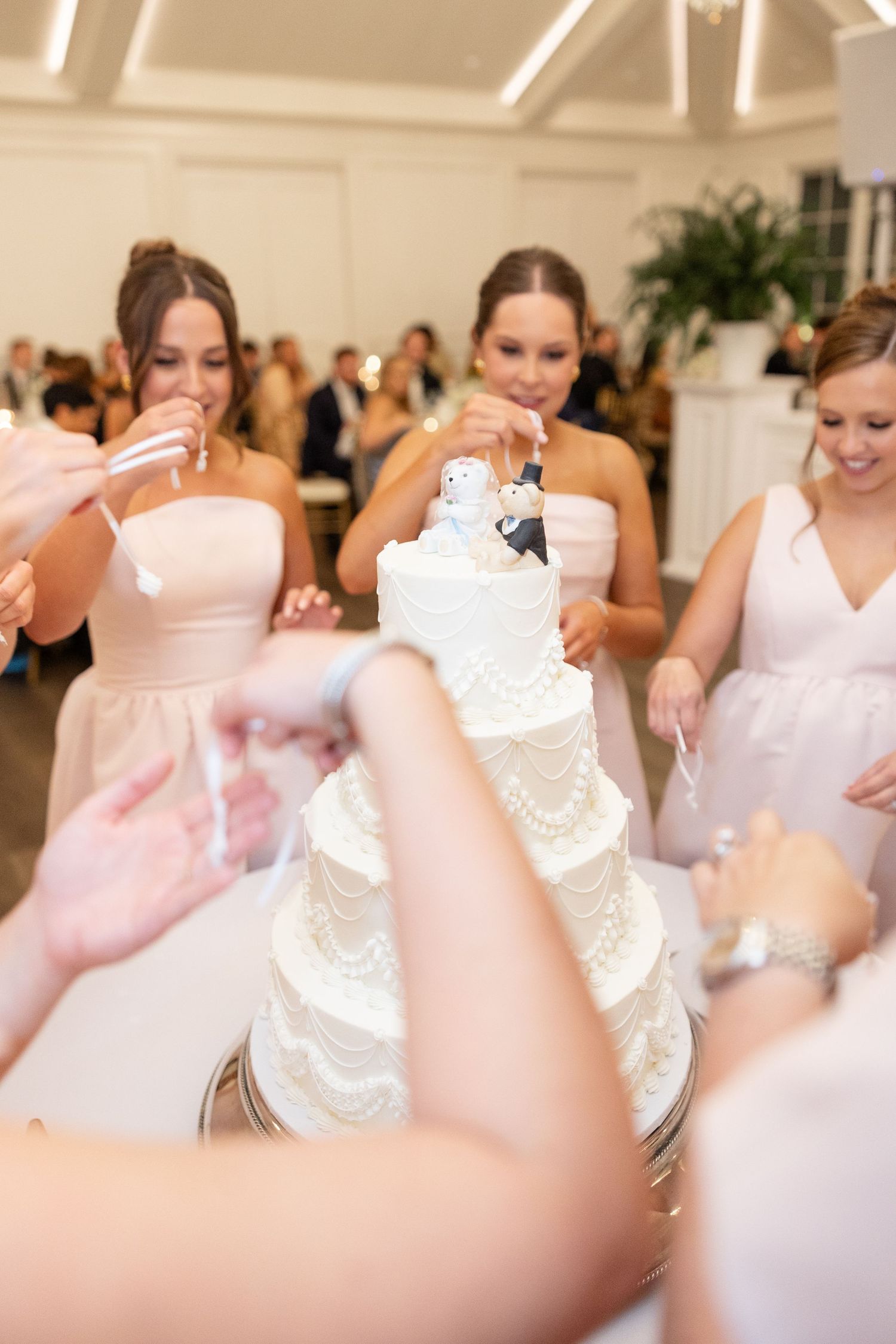 Bridesmaids in pink dresses pulling ribbons from elegant four-tier white wedding cake.