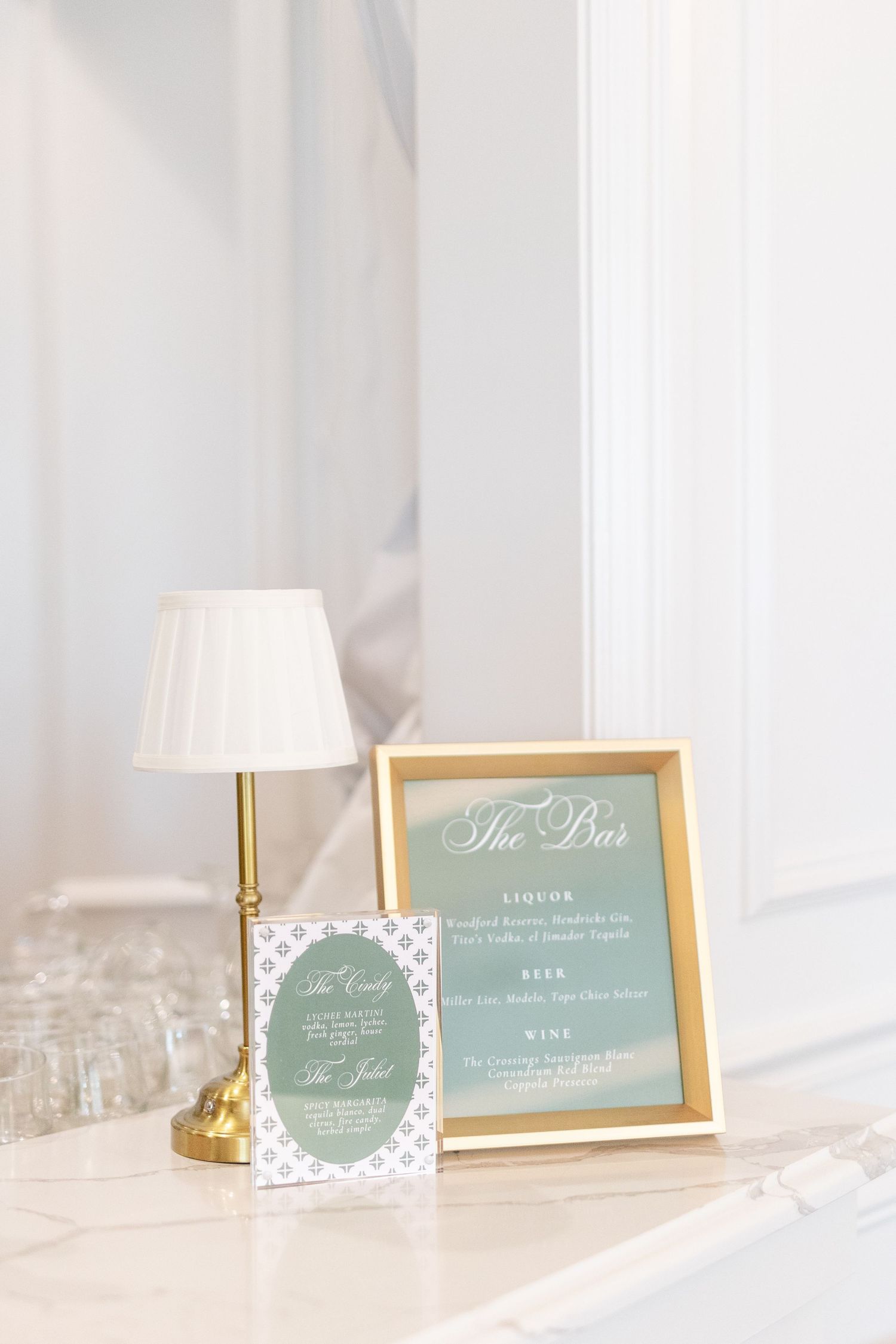 Gold-framed bar menu sign in sage green beside brass lamp and cocktail glasses at wedding reception bar.