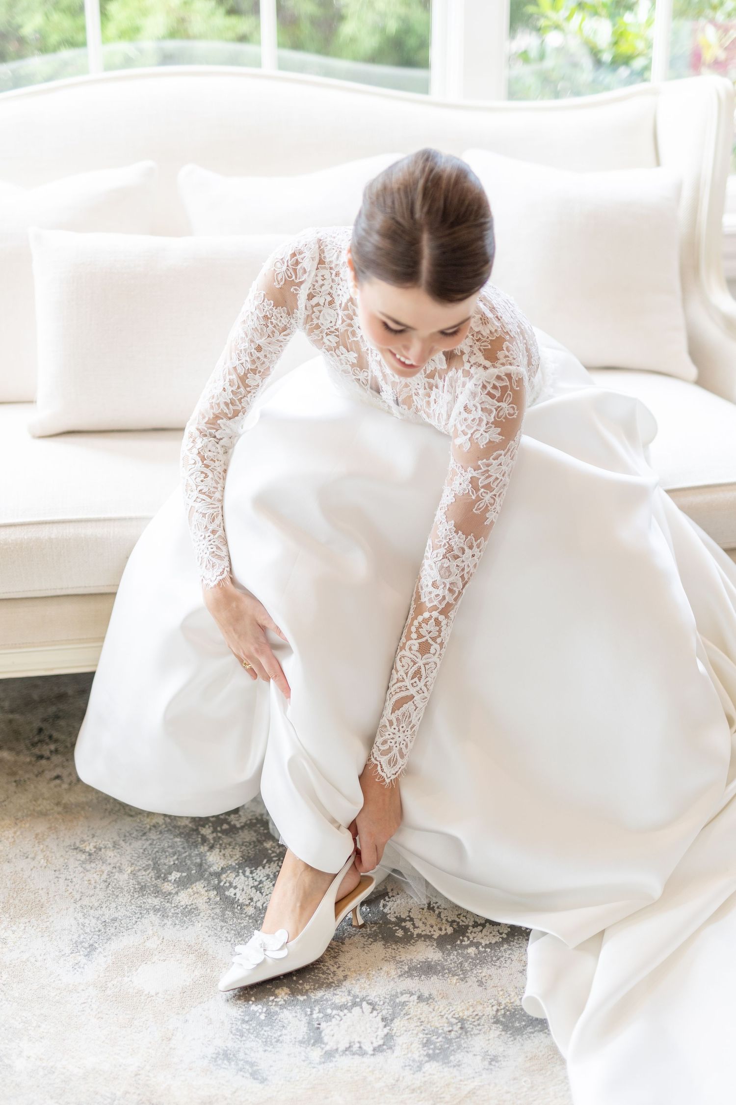 Bride in lace sleeve ball gown wedding dress putting on white heels while seated.