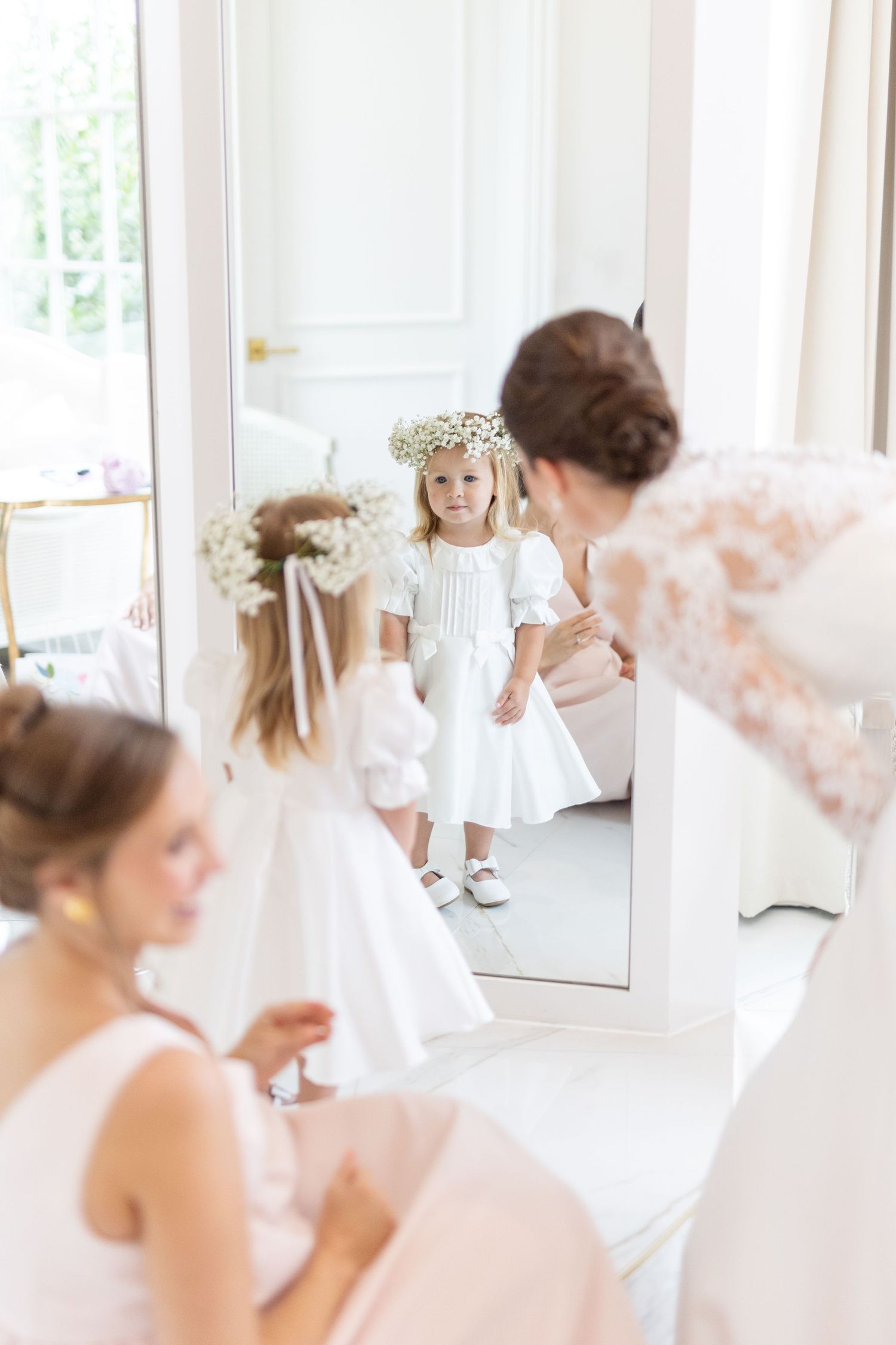 Flower girl wearing baby's breath crown smiling at her reflection in a tall mirror.