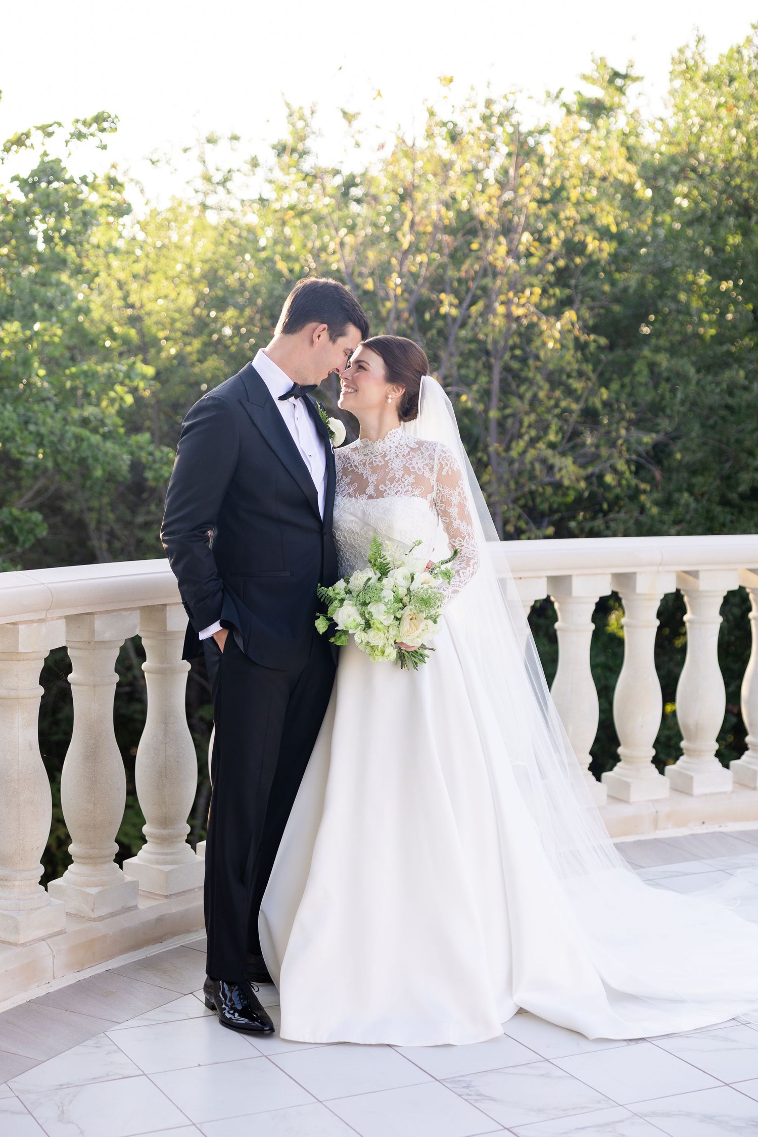 Bride and groom share a romantic forehead touch on an elegant stone balcony at golden hour.