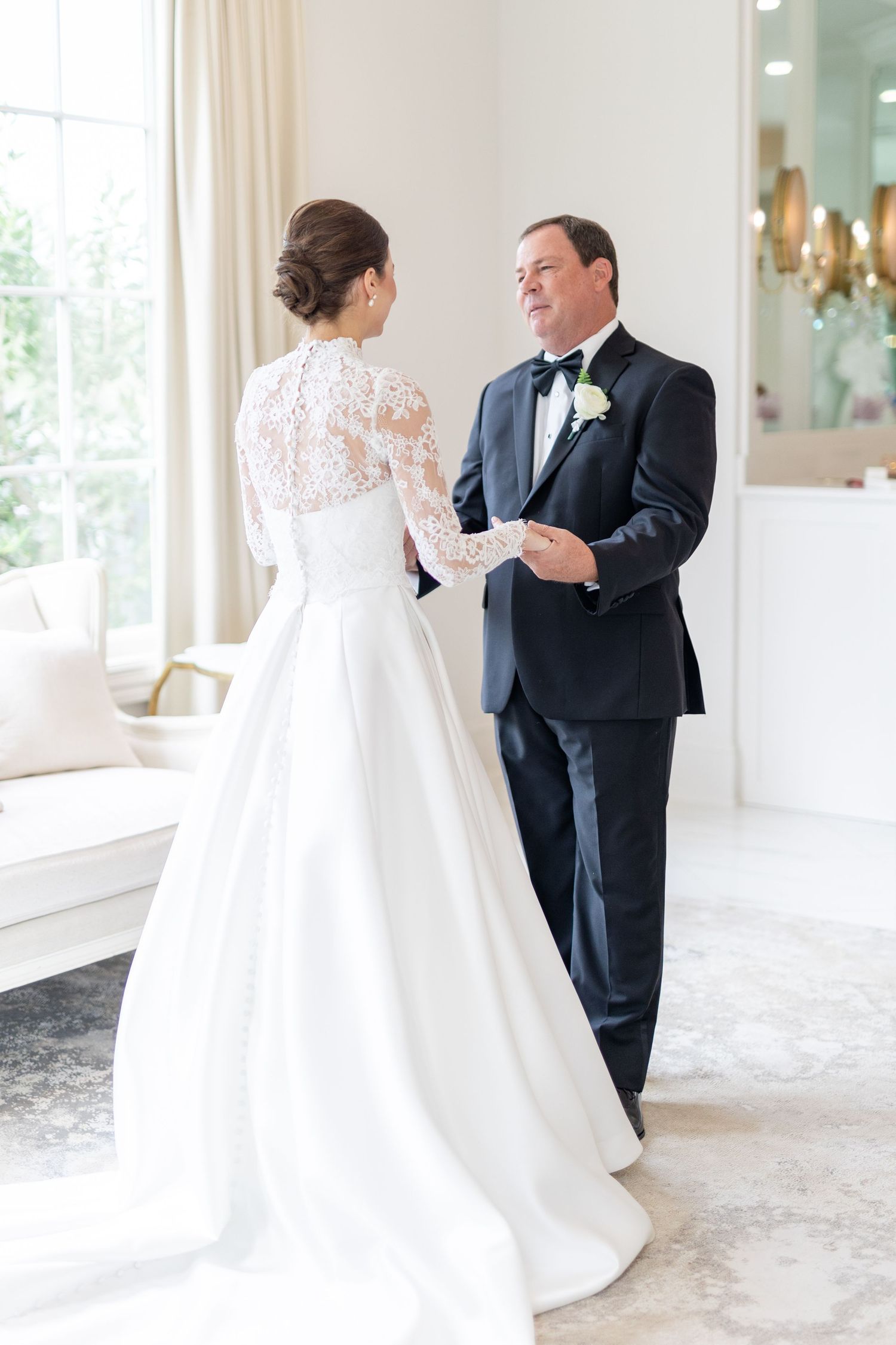 Father of the bride holds daughters hands admiring her elegant white lace wedding gown.