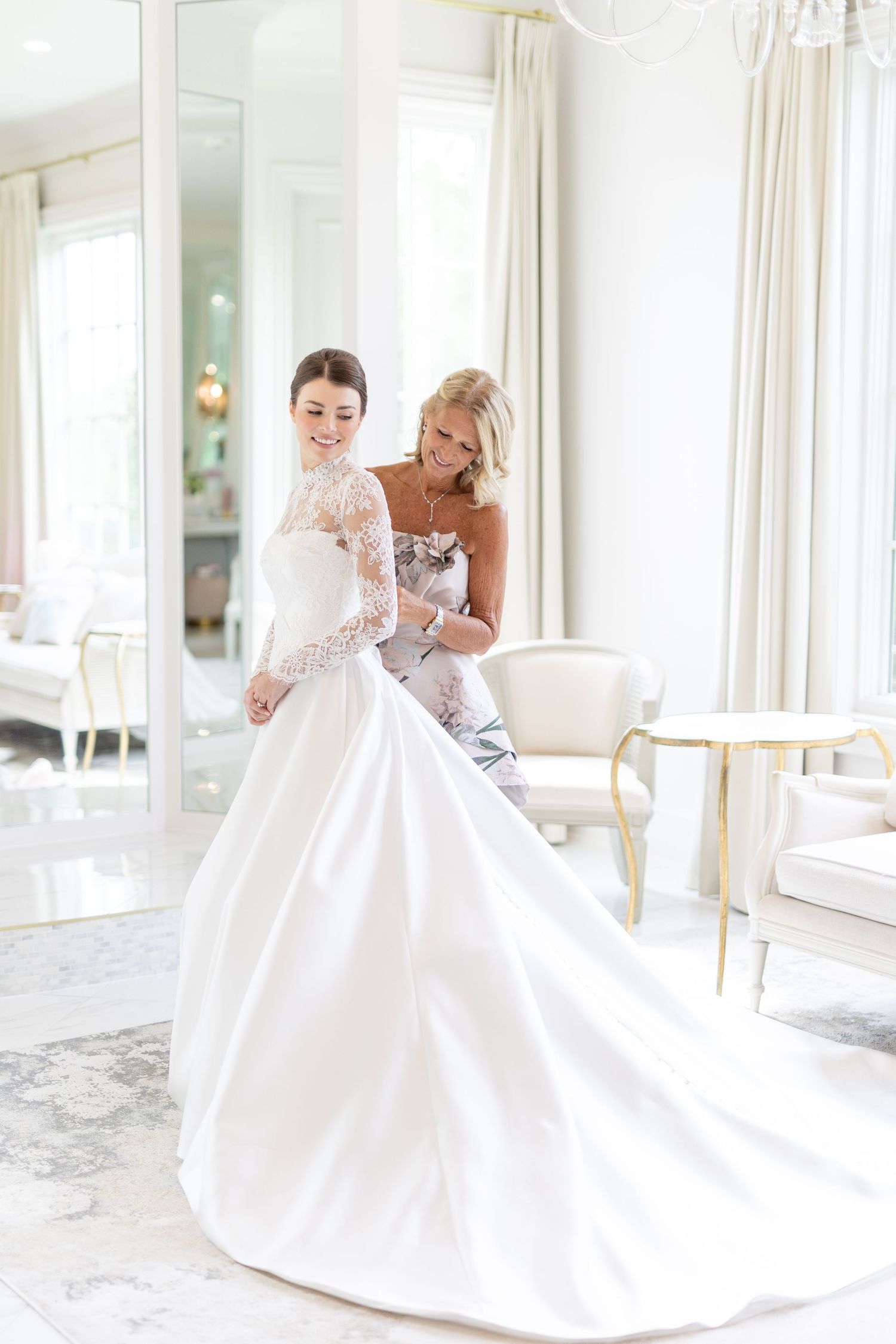 Mother helps bride into elegant white lace ball gown wedding dress in bright bridal suite.